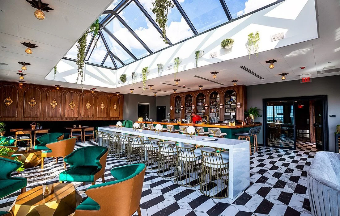 Interior of a modern bar with a large skylight, green and brown seating, a white bar counter, and a black and white checkered floor.