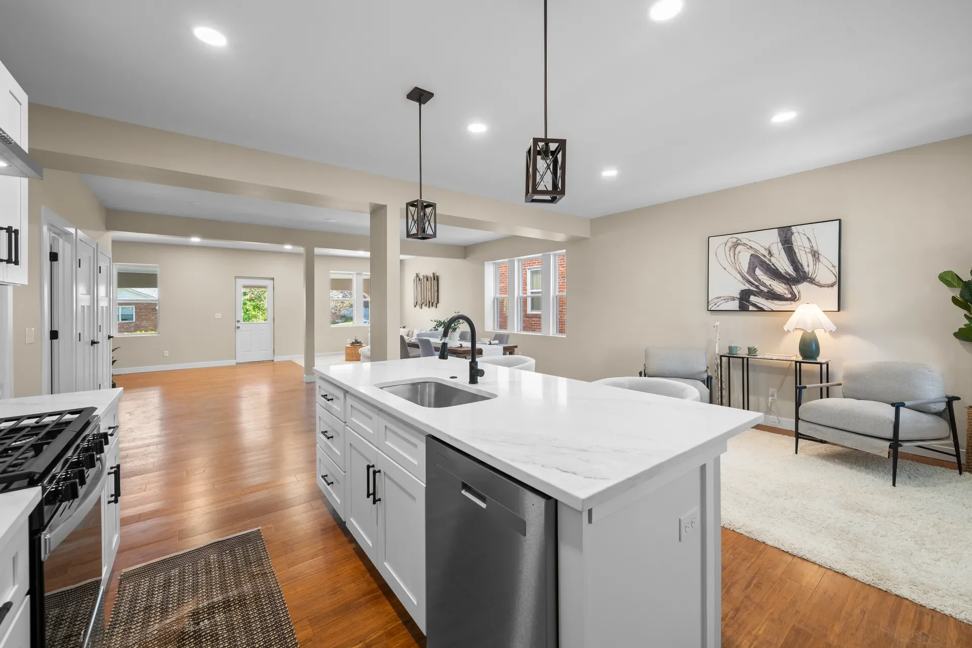 Open-concept living room and kitchen with hardwood floors, white cabinetry, a kitchen island with a sink, hanging pendant lights, beige walls, modern art on the wall, and a seating area with chairs and a side table with a lamp.