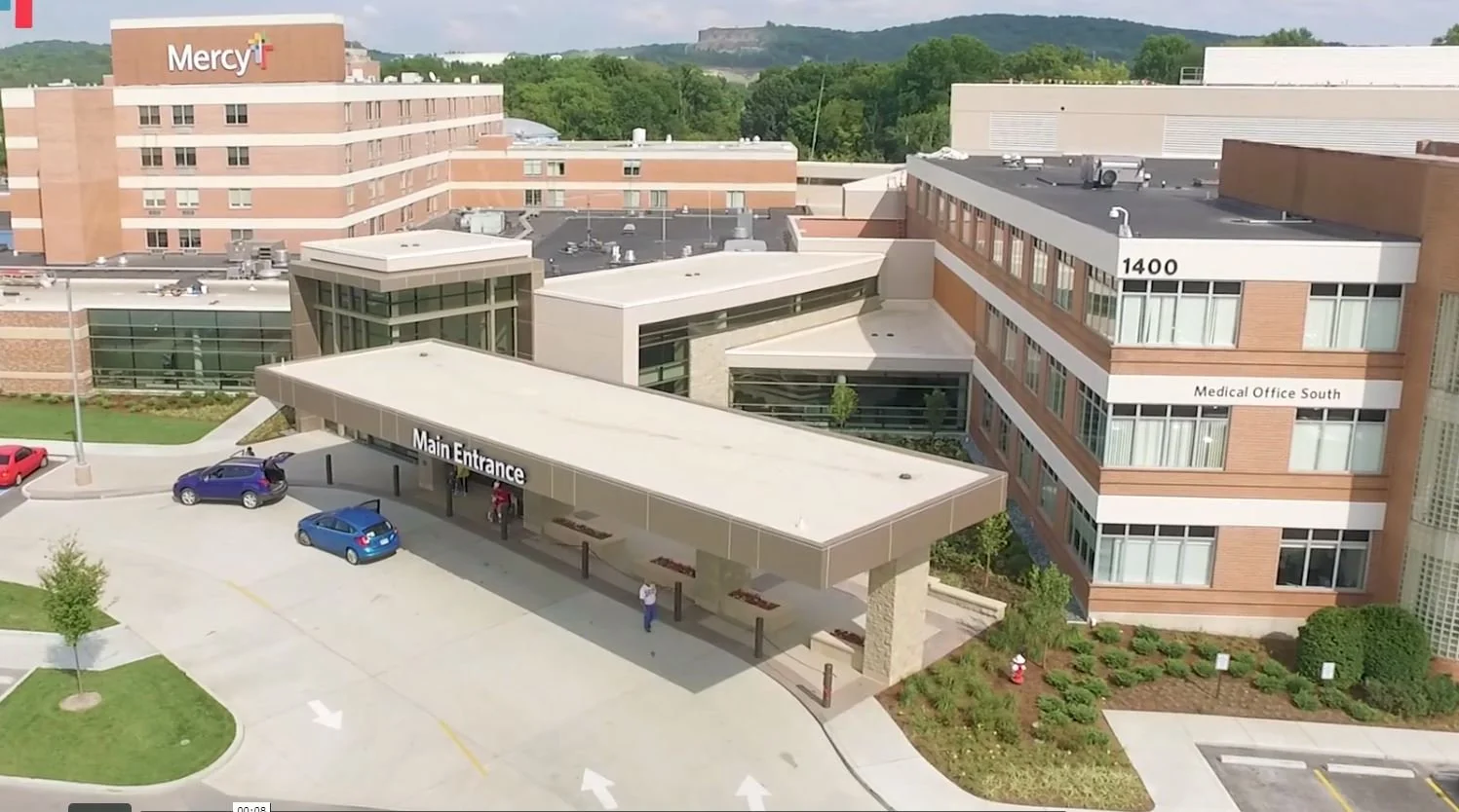 Aerial view of a hospital building with a sign for the main entrance and medical office south, parking lot with cars, and surrounding greenery.