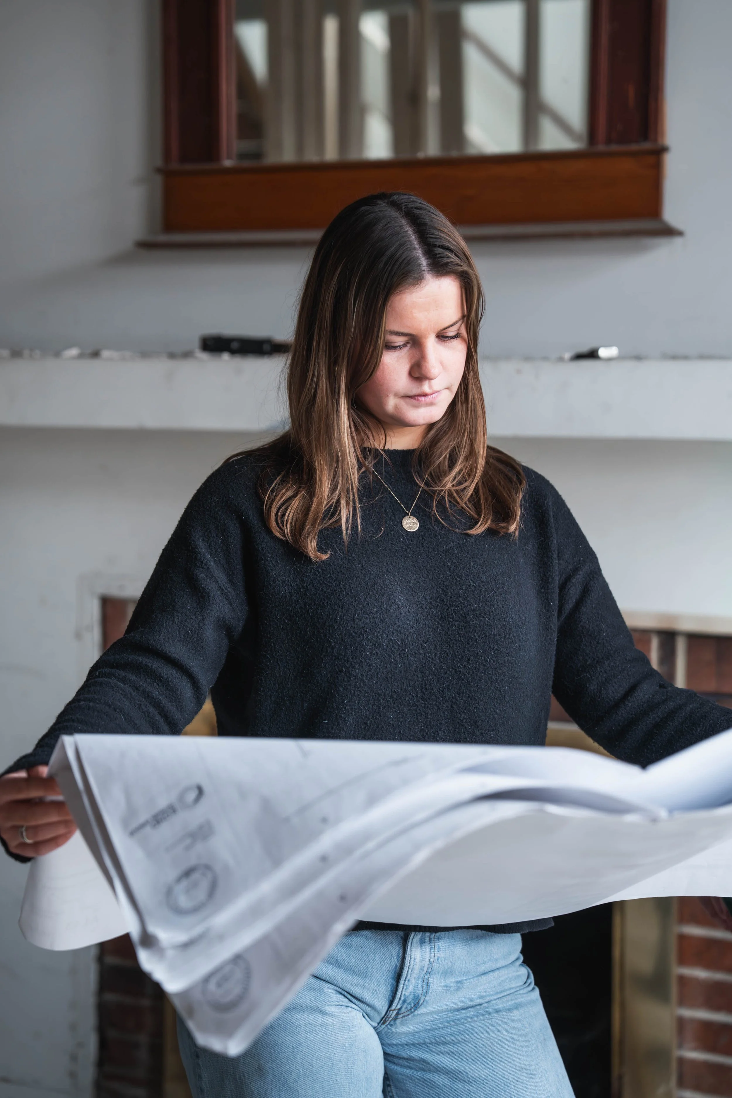 A young woman with brown hair wearing a black sweater and light blue jeans, reading a large newspaper or magazine indoors with a brick wall and window in the background.