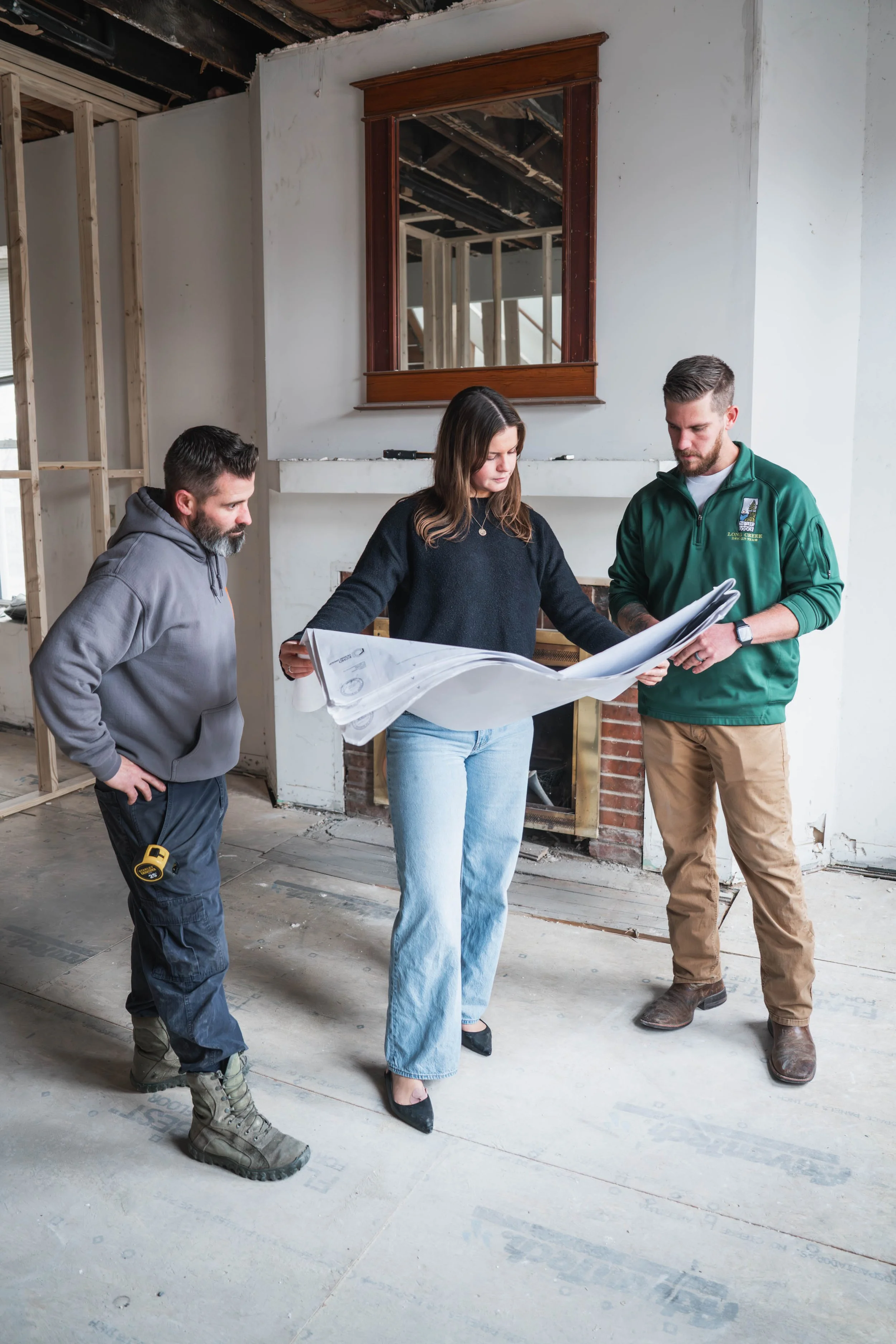 Three people, two men and one woman, are standing on a construction site looking at blueprints. The site is unfinished with visible wooden framing, an exposed ceiling, and partial wall insulation.