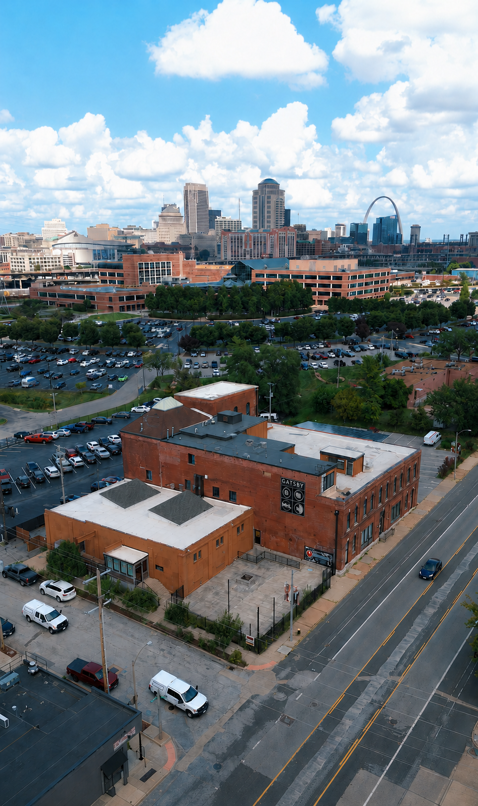 Aerial view of downtown St. Louis, Missouri, with the Gateway Arch in the background, city buildings, green trees, parking lots, and a brick building with a sign that says 'Gatsby' in the foreground.