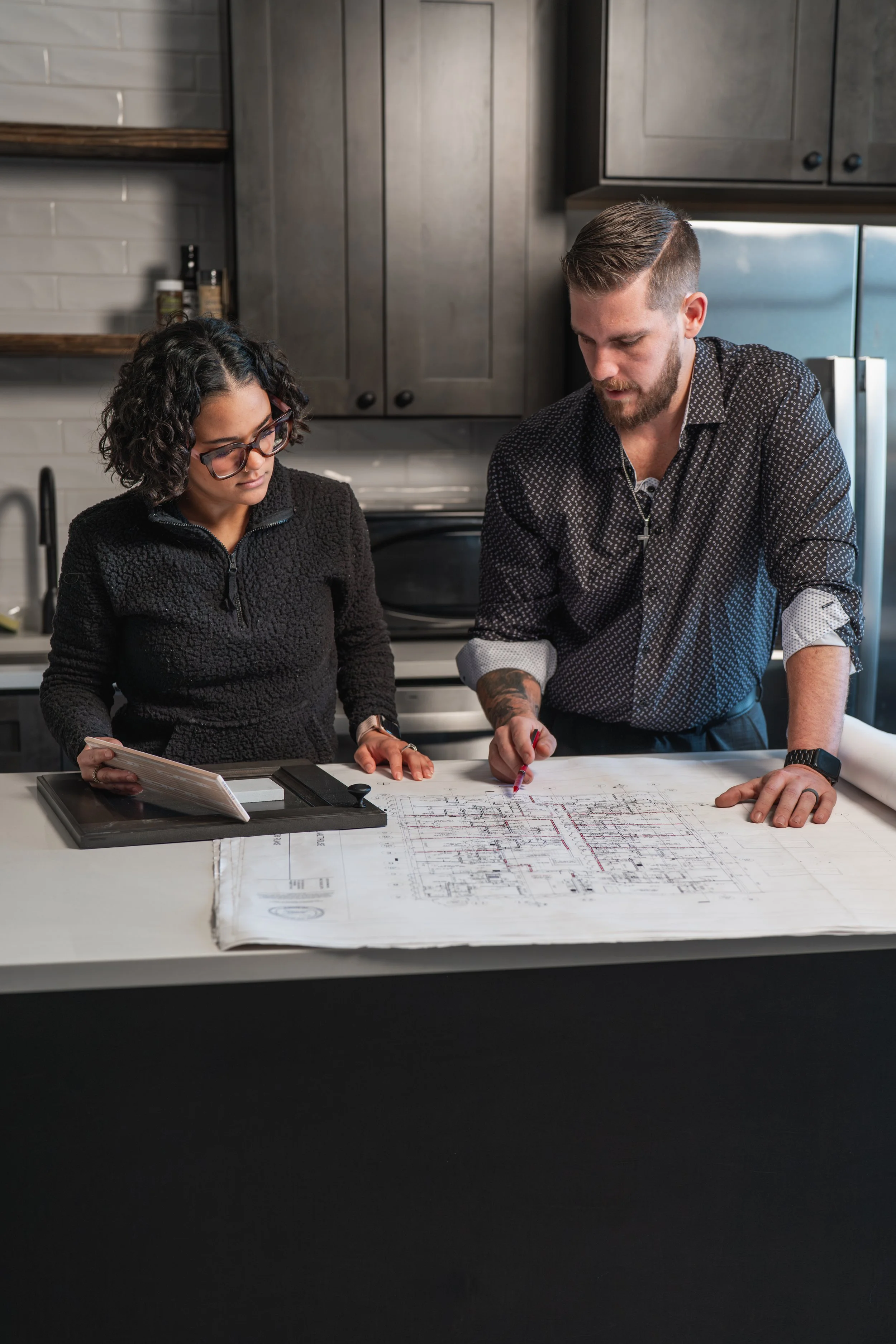 Two people, a woman and a man, working together on an architectural blueprint at a kitchen table. The woman has curly hair, glasses, and is holding a small stack of papers. The man has short hair, a beard, tattoos on his arm, and is pointing at the blueprint with a red pen. They are focused on the plan in a modern kitchen setting.