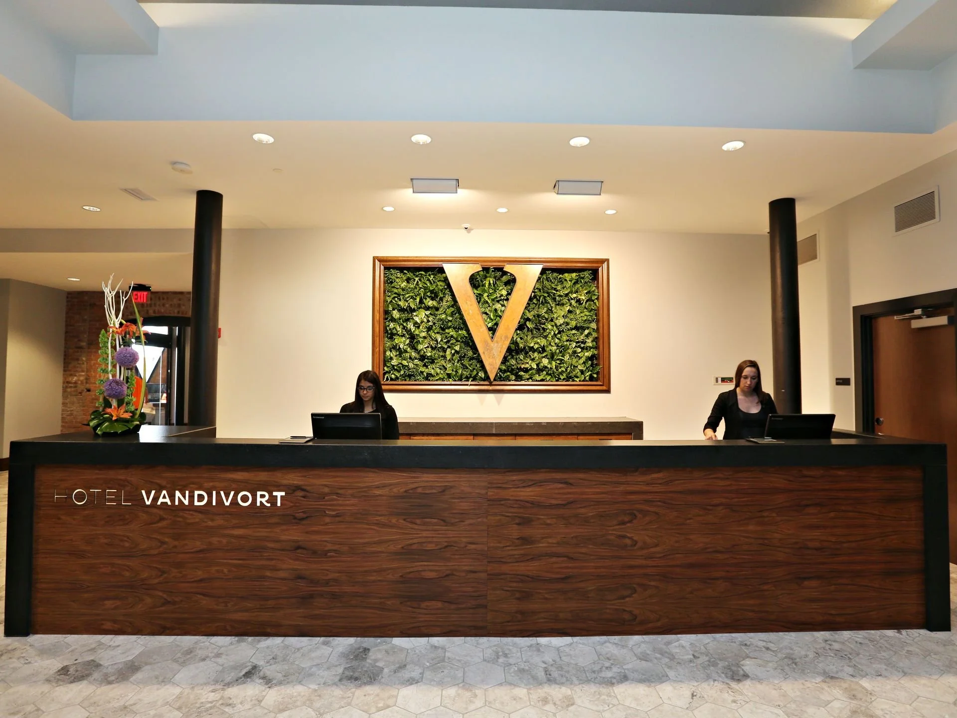 Hotel lobby reception desk at Hotel Vandivort with a wooden front desk, a large gold V logo on a greenery background on the wall behind the desk, and two staff members working at computers.
