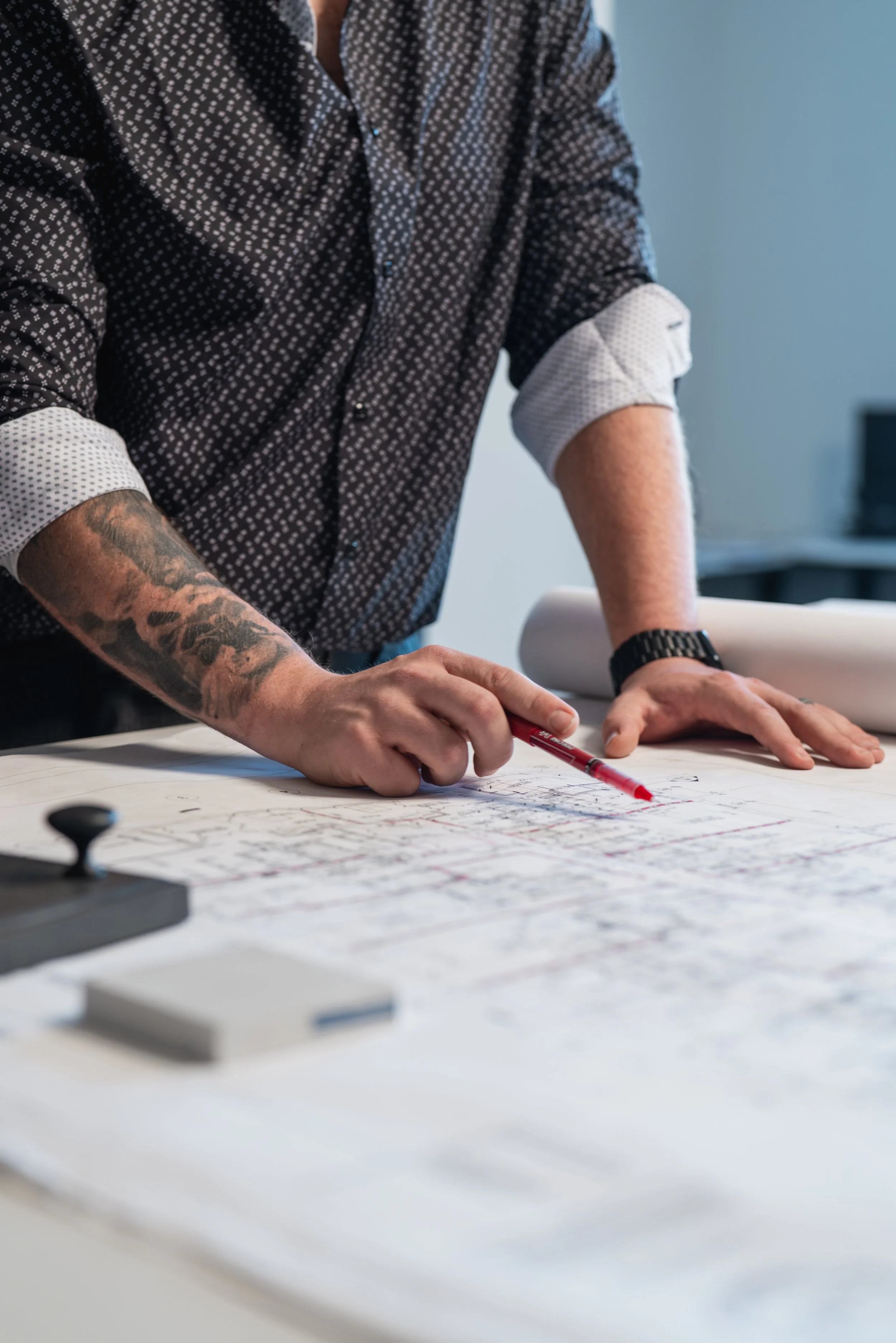 A person with tattooed arms wearing a black patterned shirt with rolled-up white sleeves is writing or drawing on large sheets of paper on a table with a red marker. In the background, there's a blurred desk with a computer.