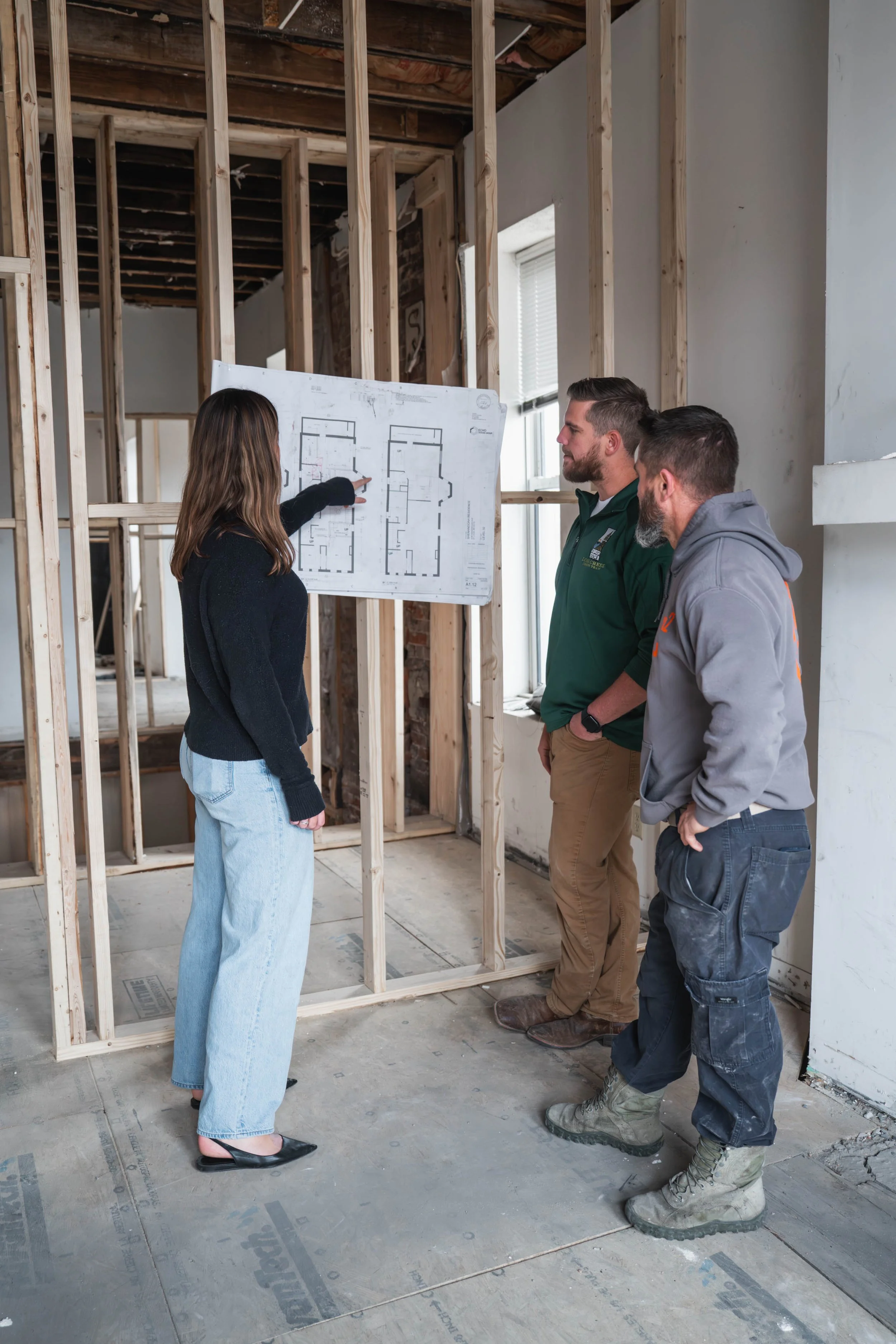 Three people, a woman and two men, are discussing a floor plan on a construction site with exposed wooden framing and unfinished walls.