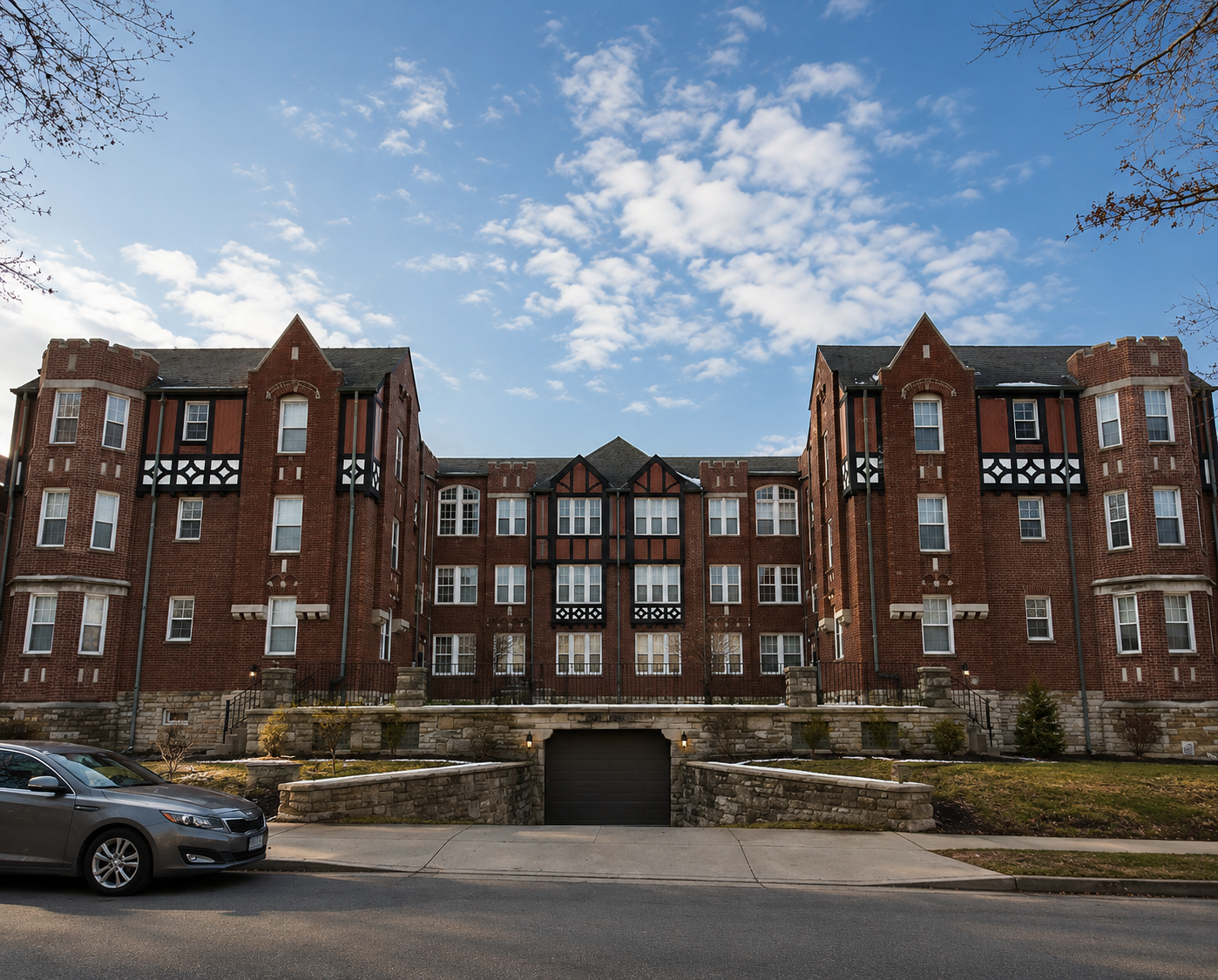 A multi-story red brick apartment building with black and white decorative trim, shaped like a castle, under a partly cloudy sky. A parked silver car and an entrance to an underground garage are visible in the foreground.