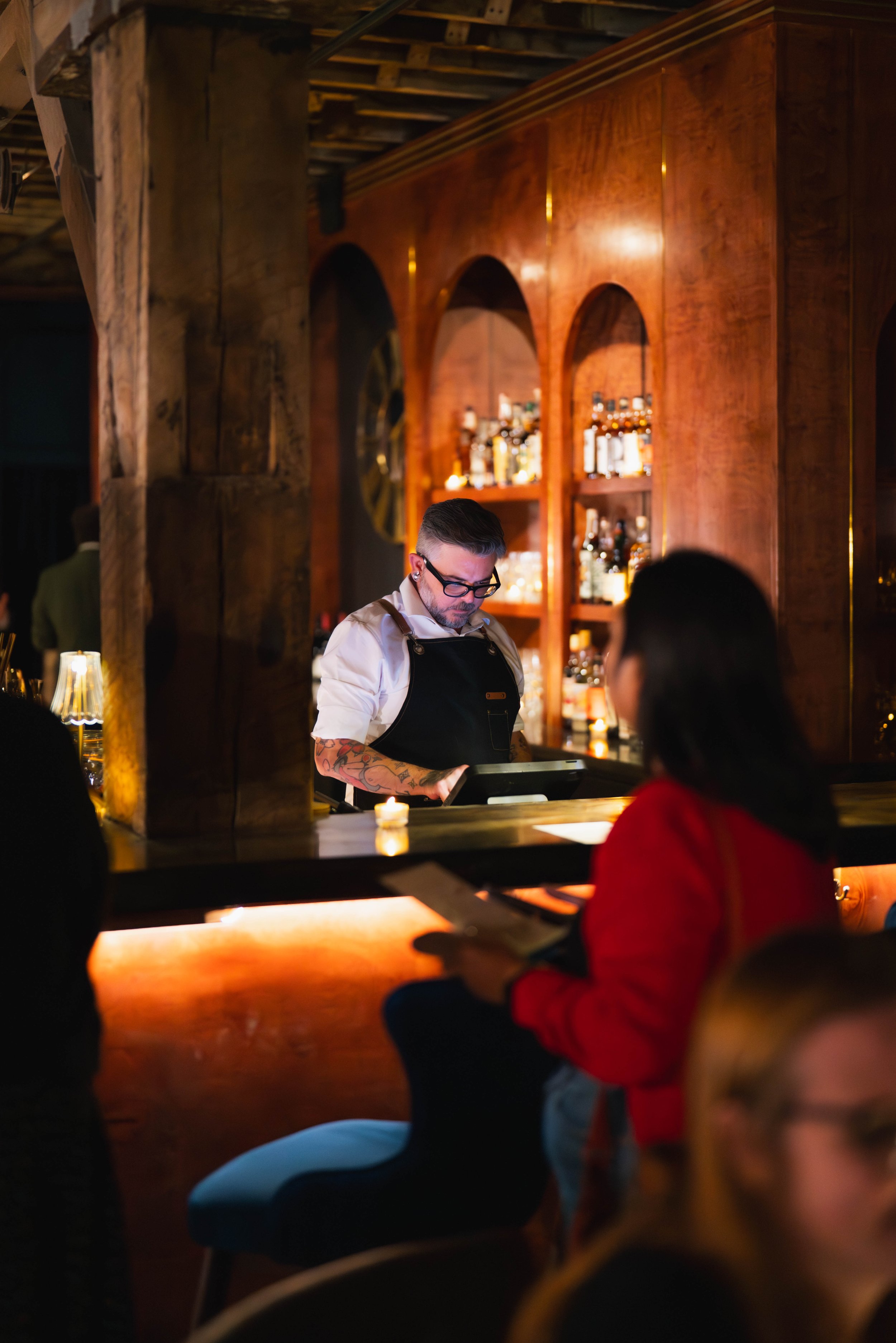 A bartender with glasses and tattoos on his arm looking at a tablet in a dimly lit bar with wooden shelves and bottles in the background. There are patrons in the foreground, one in a red shirt.