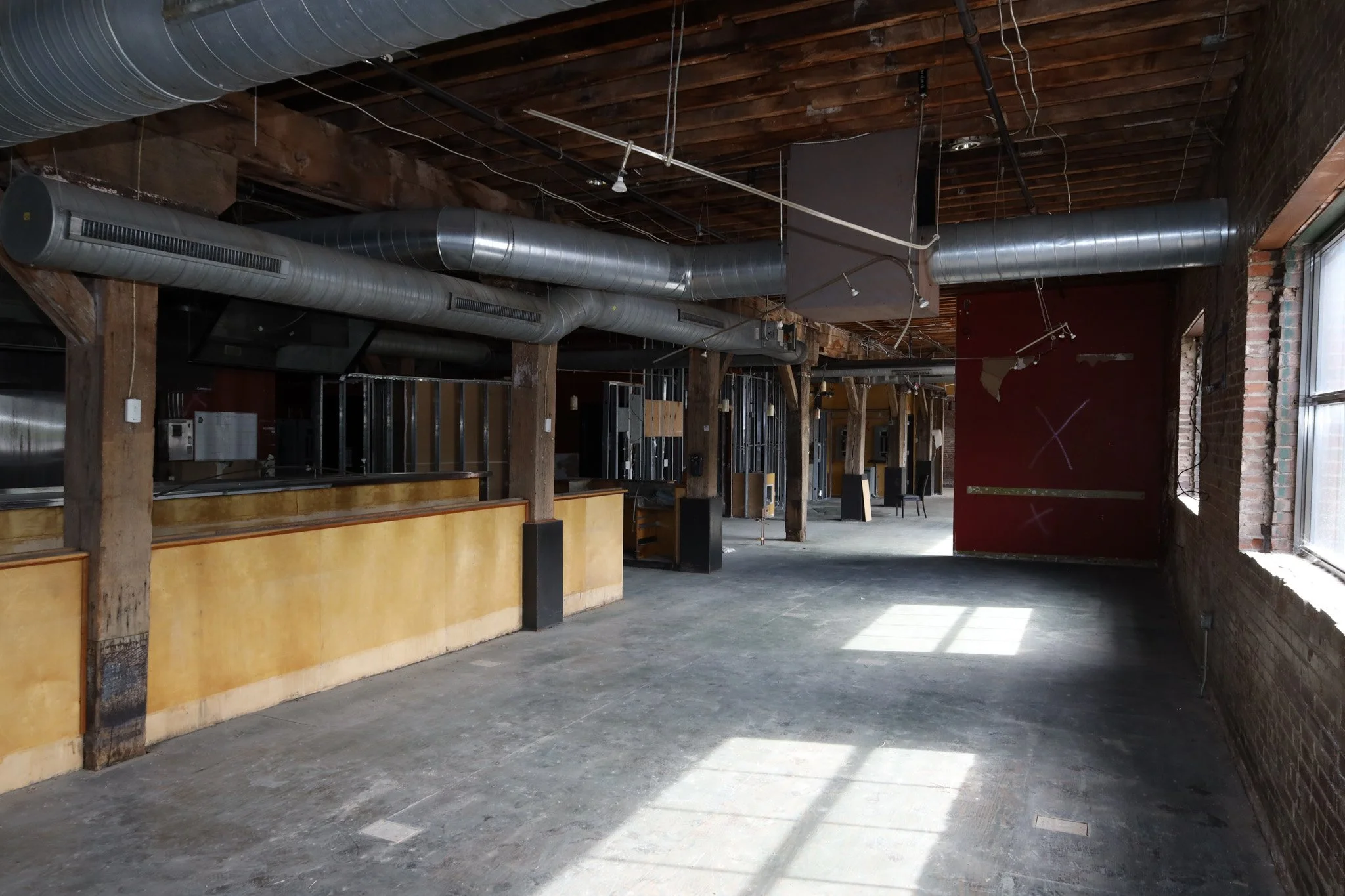 Empty interior space with exposed ductwork, wooden beams, brick walls, and large windows letting in natural light.