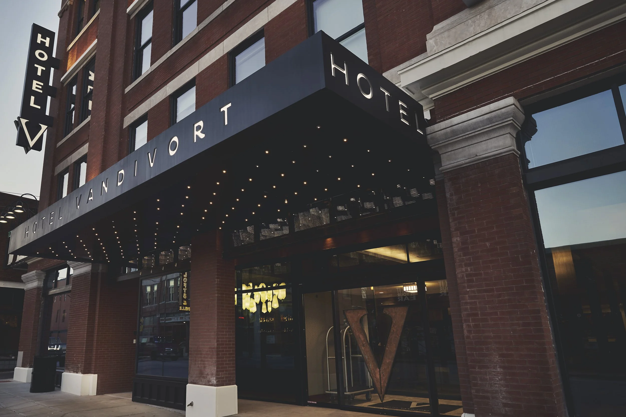 Exterior view of a brick hotel building with a black awning, illuminated with small lights, displaying the words 'Landvor Hotel' and enclosed within black frames.