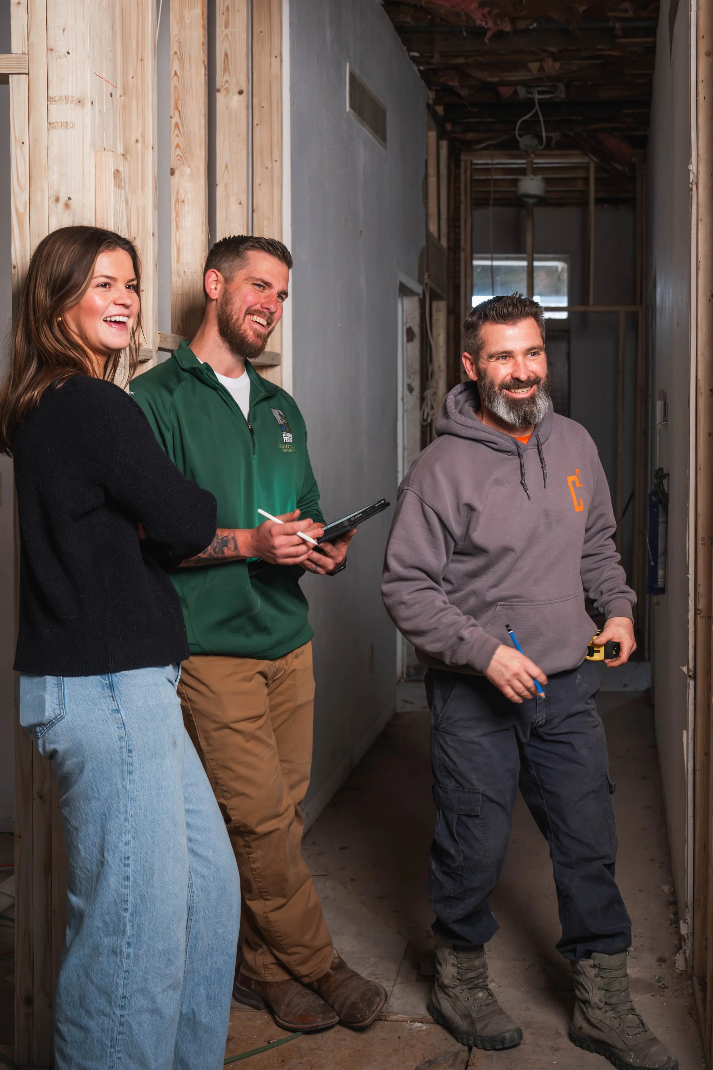 Three people standing inside a house under construction or renovation, with visible wooden framing and exposed ceiling, smiling and engaging in conversation.