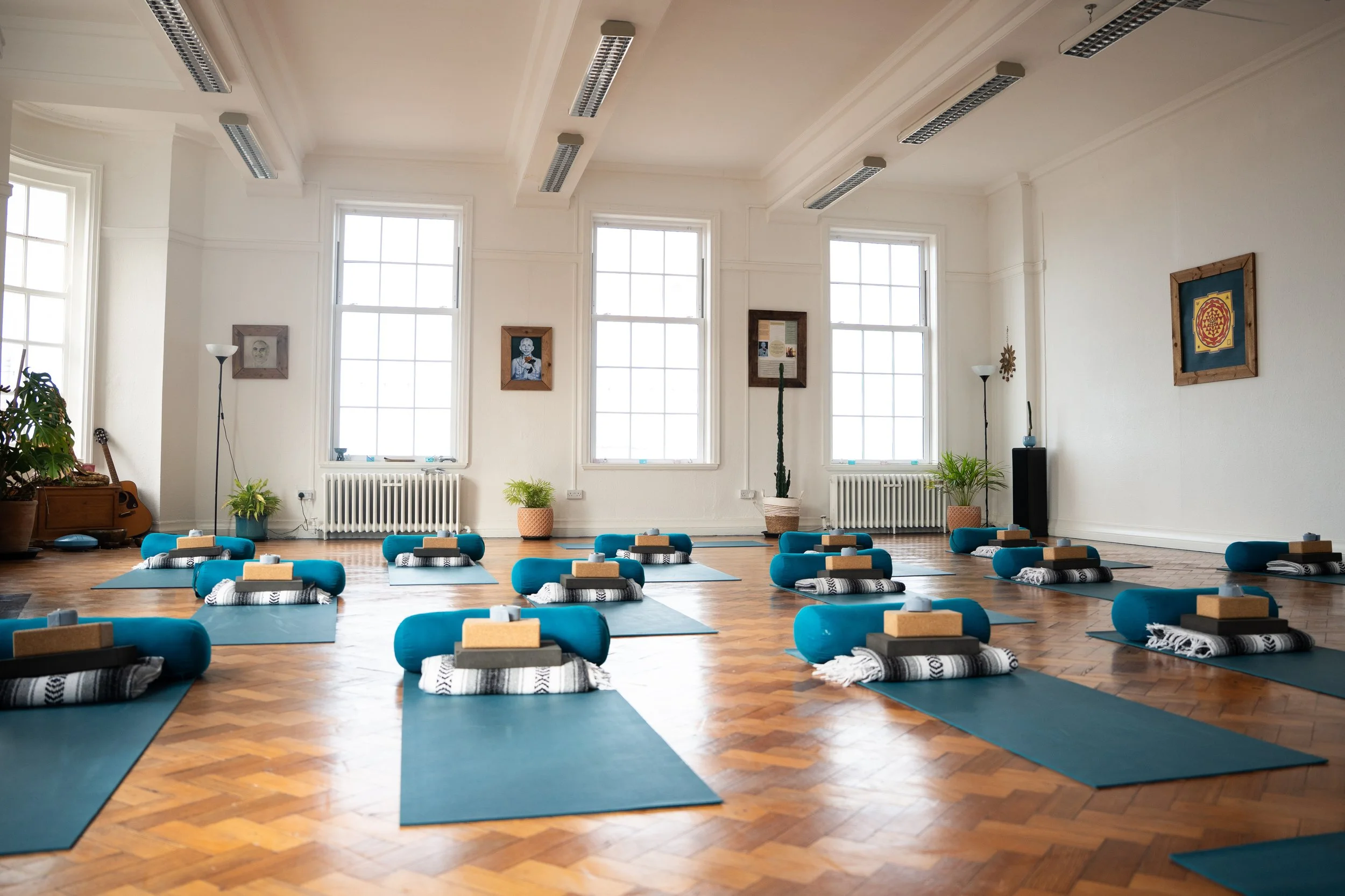 A spacious yoga or meditation room with blue mats and bolsters arranged on a wooden floor, tall windows letting in natural light, and framed artwork on the white walls.
