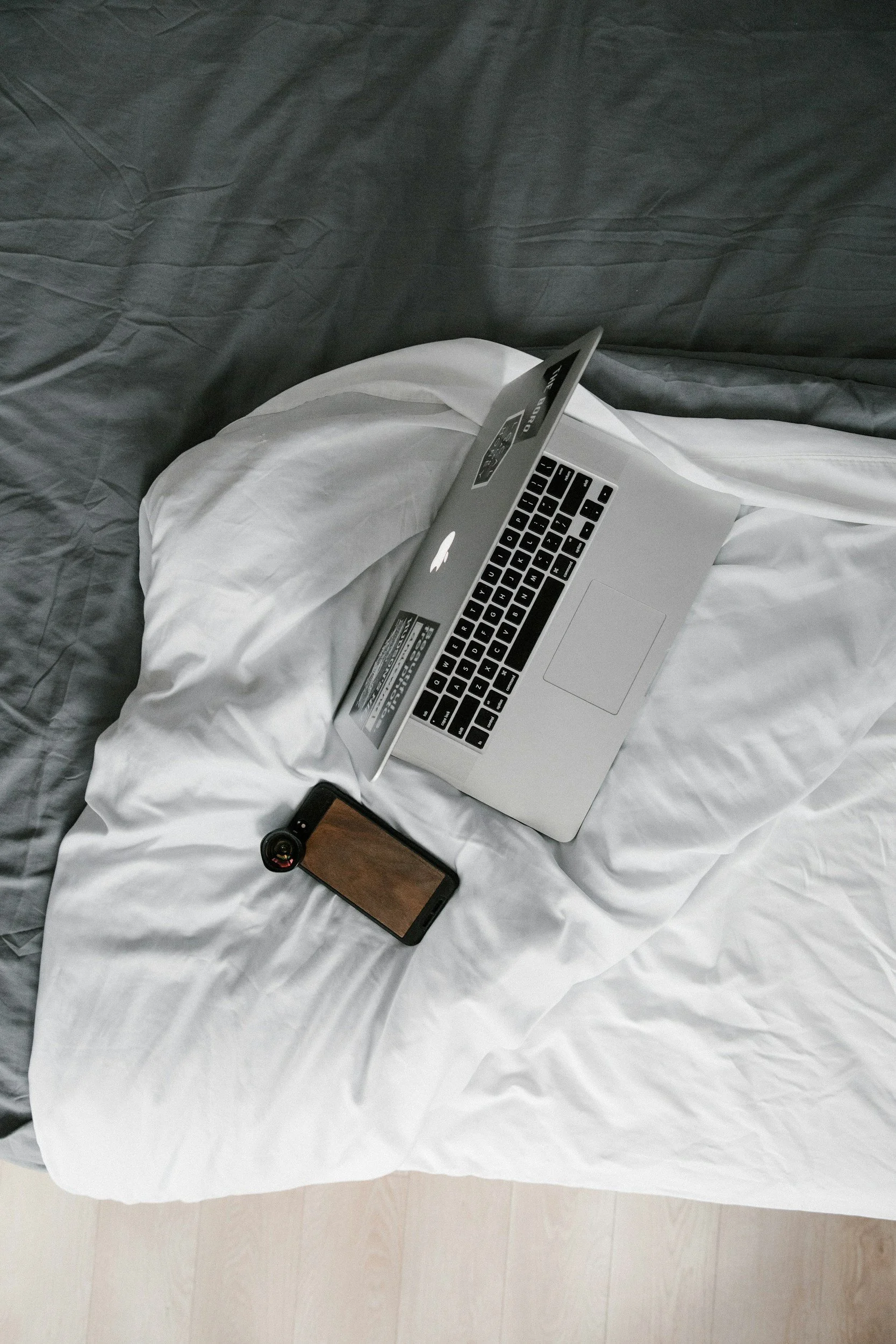 A bed with white sheets and pillows, displaying a silver MacBook laptop, a smartphone with a case, and a small camera on it.