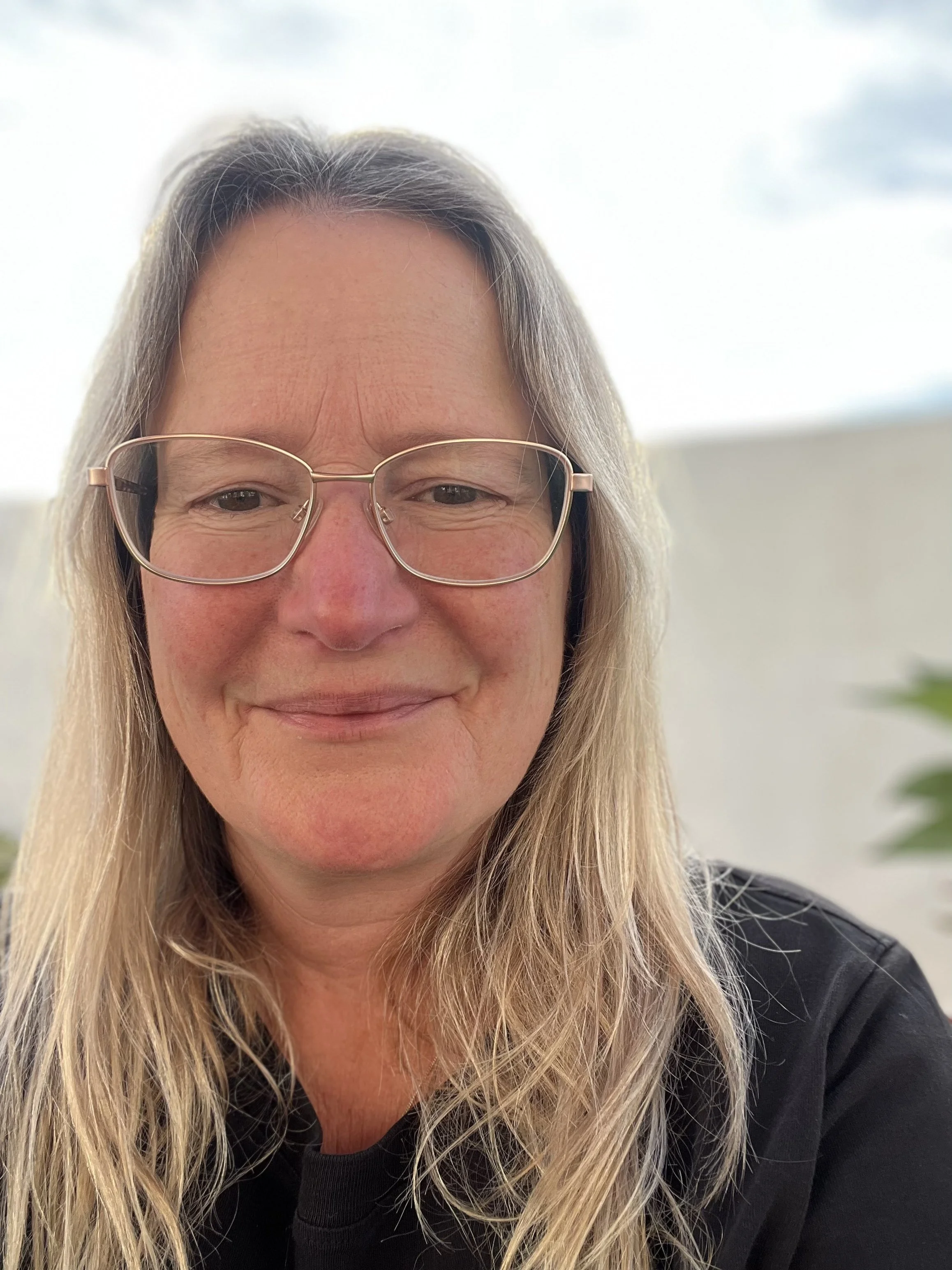 A woman with long blonde hair and glasses smiling in natural light, wearing a black shirt.