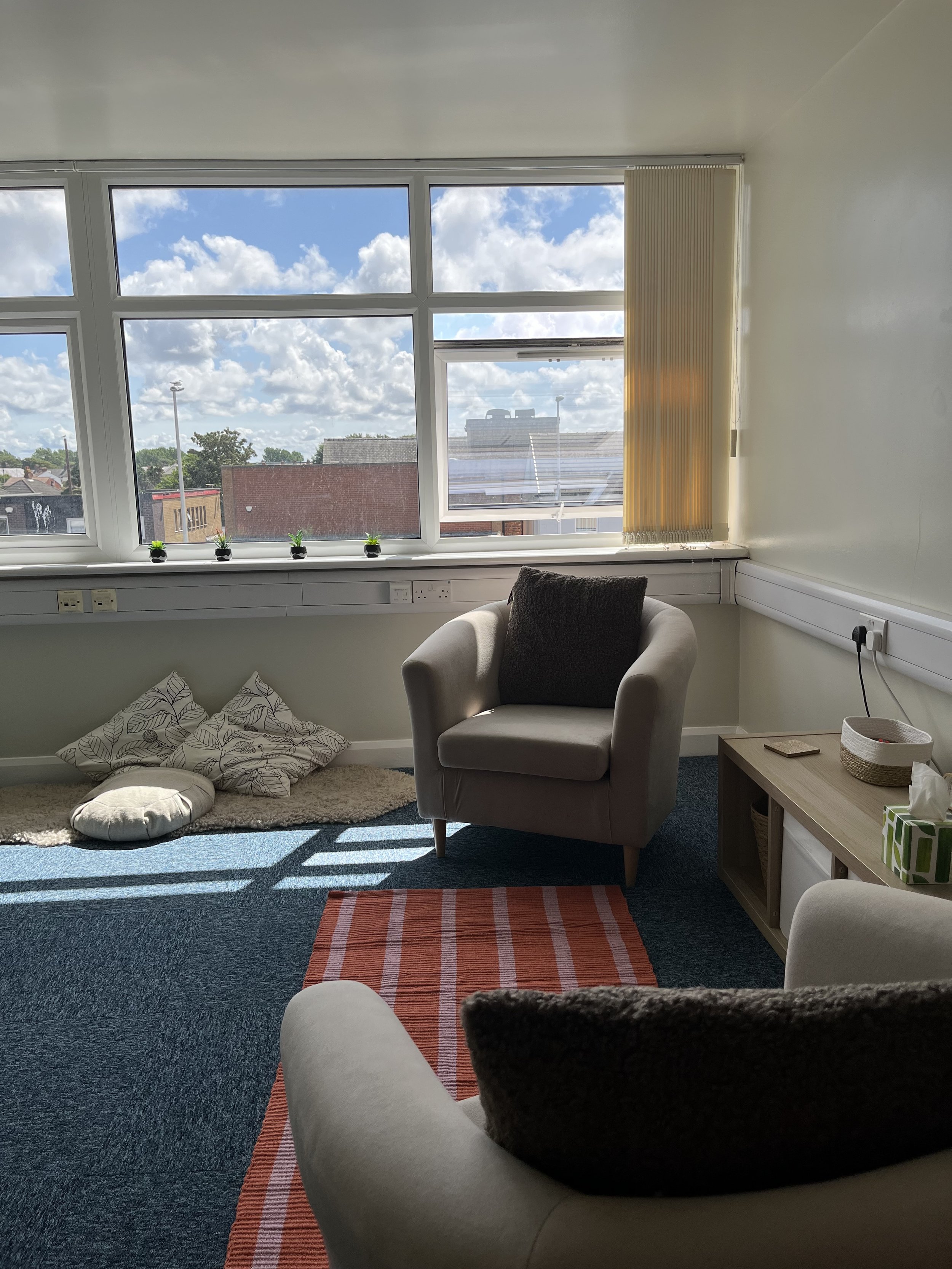 A cozy room with large front windows showing a partly cloudy sky and surrounding buildings. There are four small potted plants on the windowsill, a beige armchair with a dark cushion, a small side table with a basket and tissue box, cushions on the floor, a striped rug, and a blue carpet.