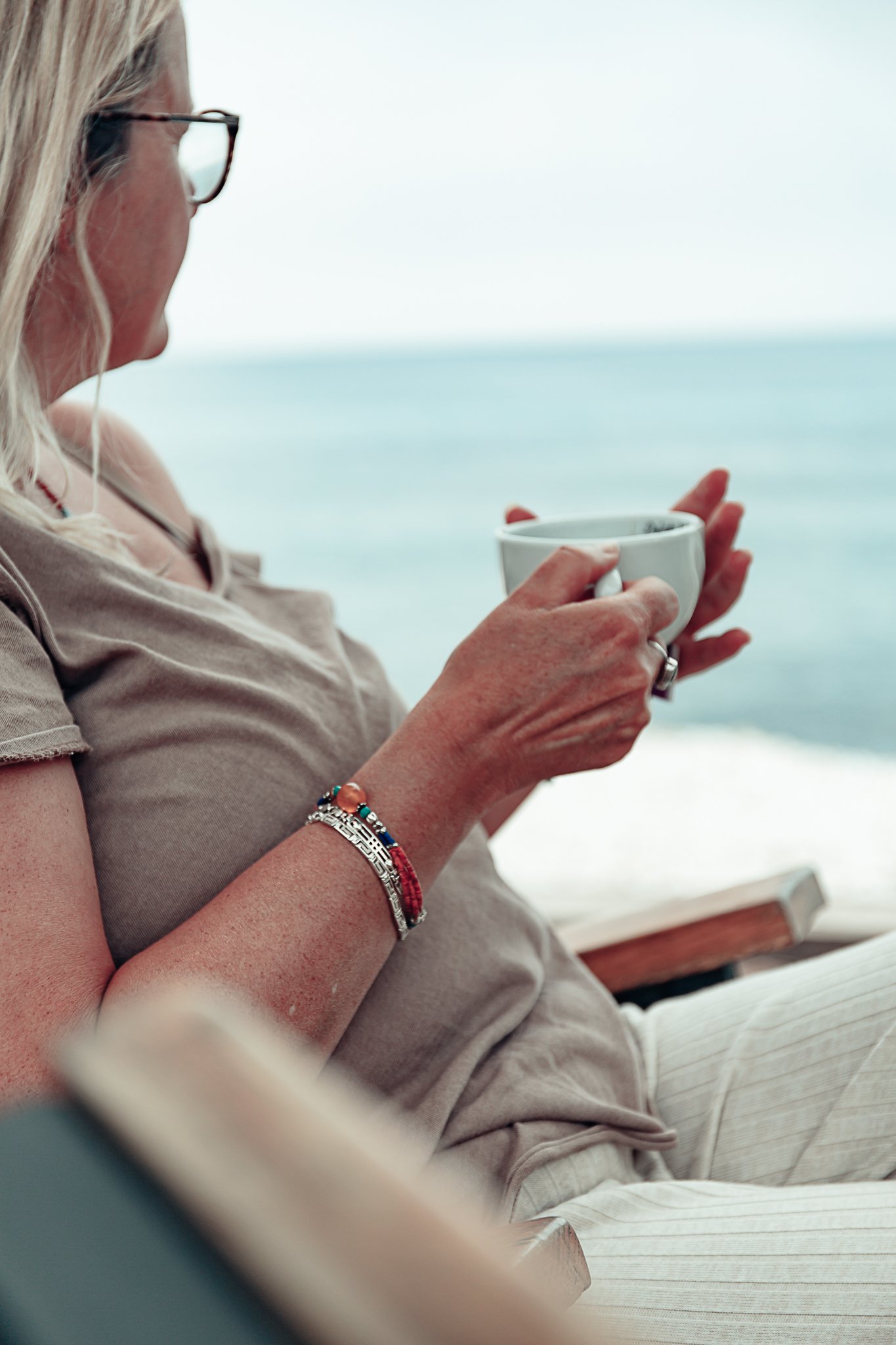 A woman with blonde hair wearing glasses and a beige top, holding a white mug, sitting outdoors with a blurred ocean background.