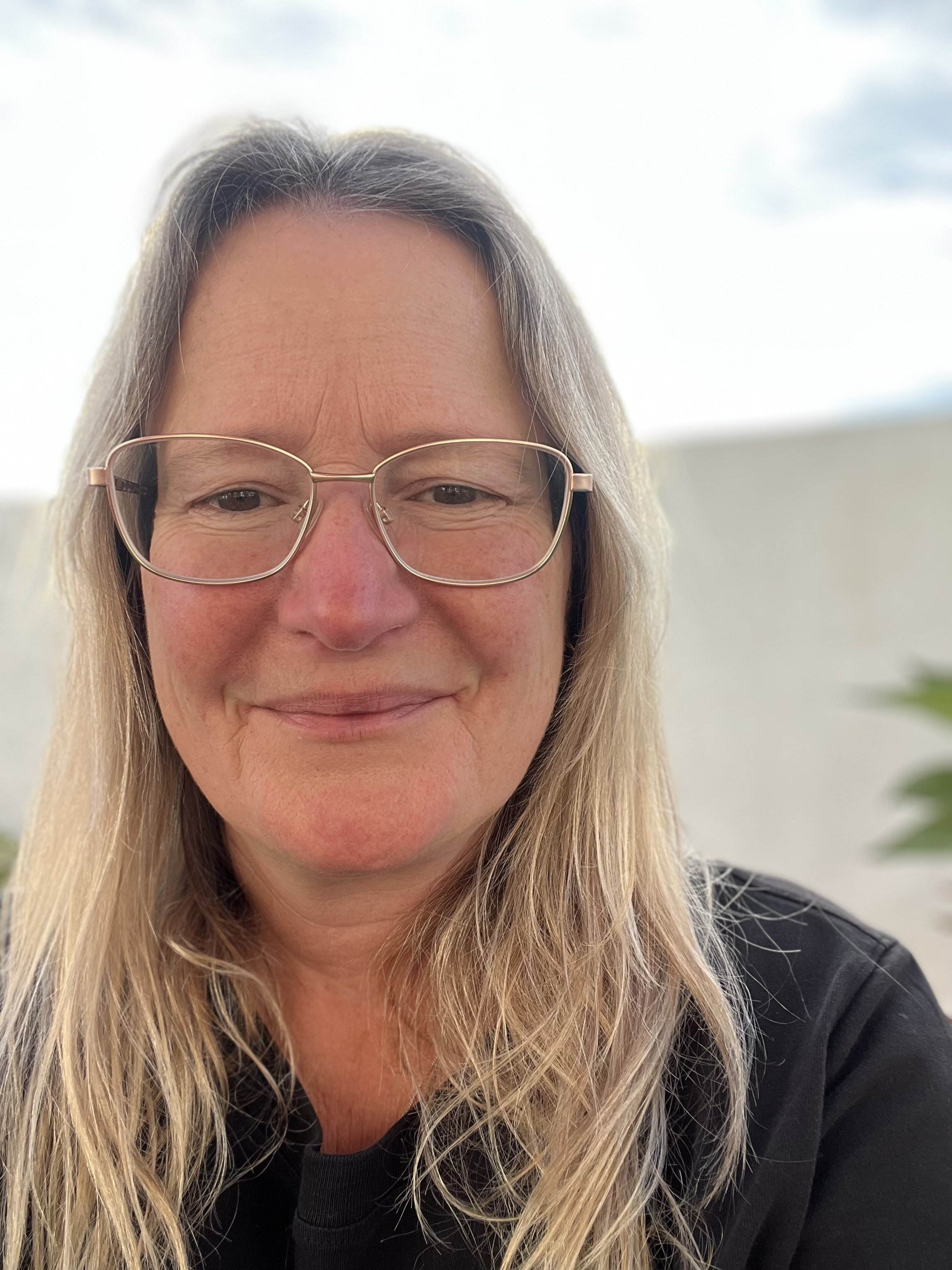 A woman with long blonde hair, glasses, and a black shirt, smiling indoors with a blurred background and a hint of a plant.
