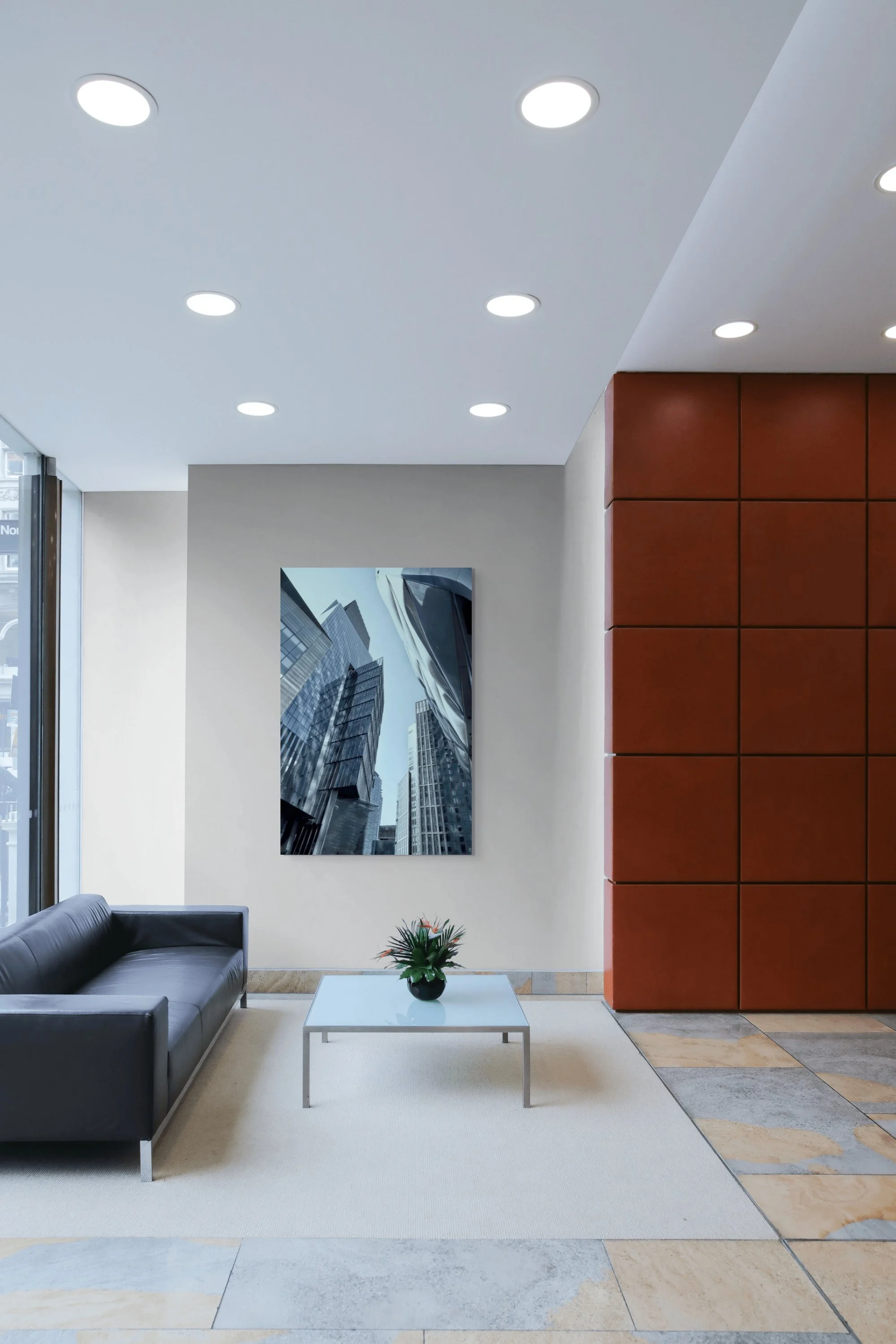 Modern office lobby with a gray couch, white coffee table with a potted plant, a large cityscape photo on the beige wall, a red tiled wall, and a partial view of a glass door and window. Recessed ceiling lights illuminate the space.