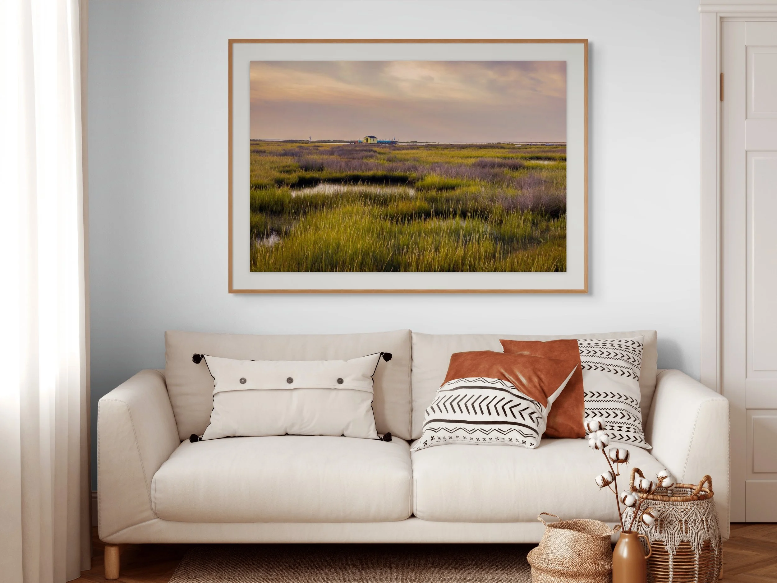 A gallery wall features a large landscape painting of marshland with grasses and a small building, hung above a cream-colored sofa with decorative pillows, in a well-lit living room.