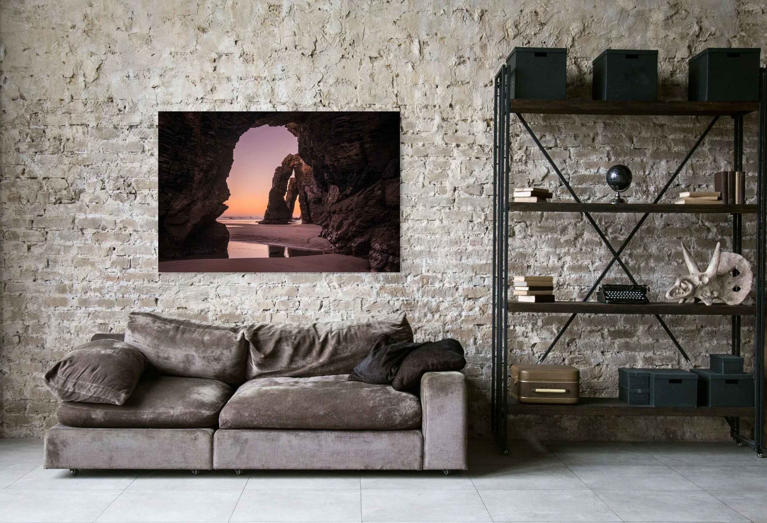 Living room with white brick wall, a gray sofa with three pillows, a black metal bookshelf with books and decorative items, and a framed sunset beach scene on the wall.