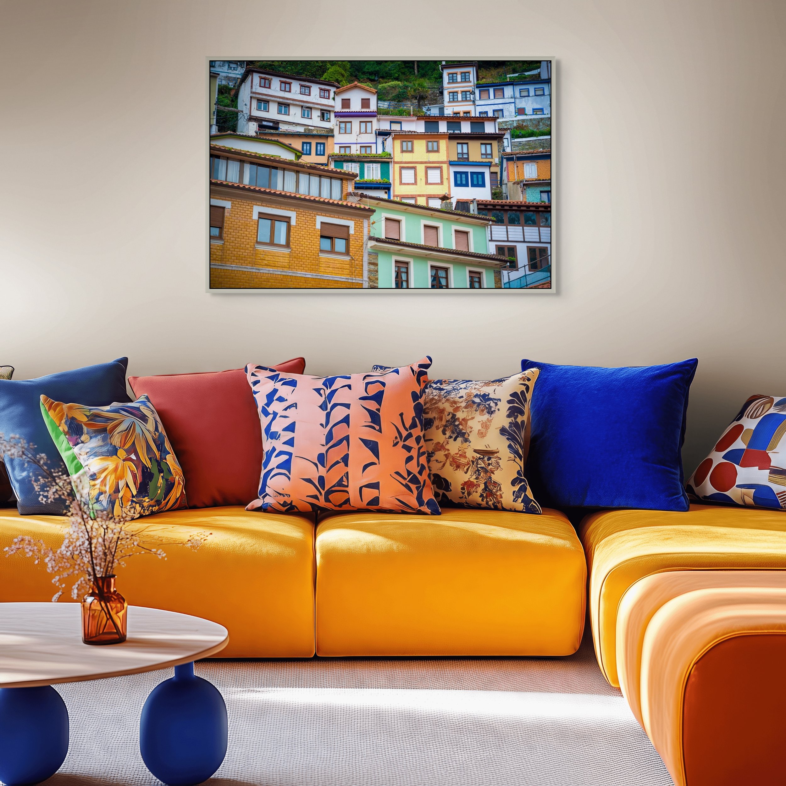 Colorful living room with yellow sofa, various patterned pillows, and a white coffee table holding a small orange glass vase with flowers. A large framed photograph of colorful hillside houses hangs on the wall.