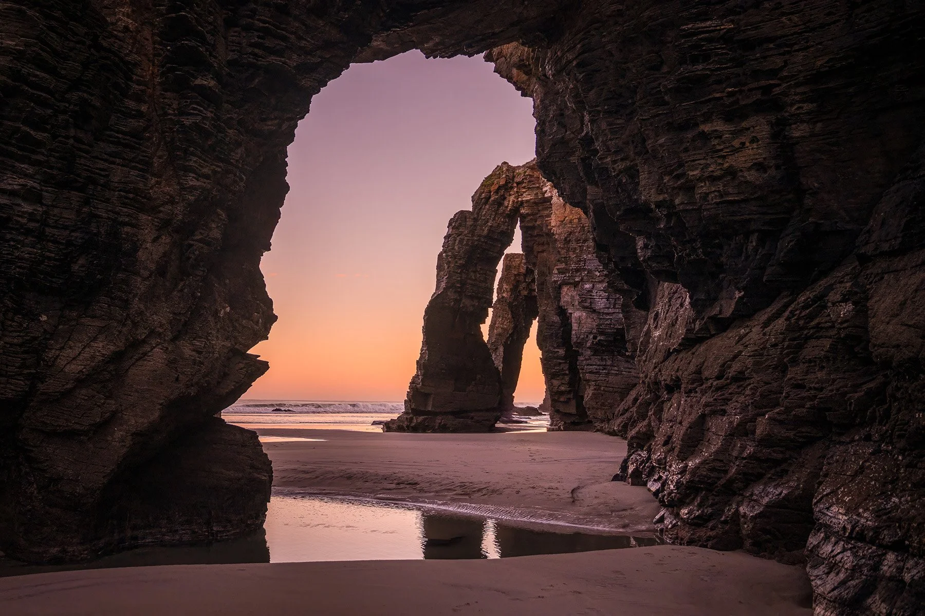 View of a beach from inside a rocky cave, with large cascading sandstone arches in the background during sunrise.