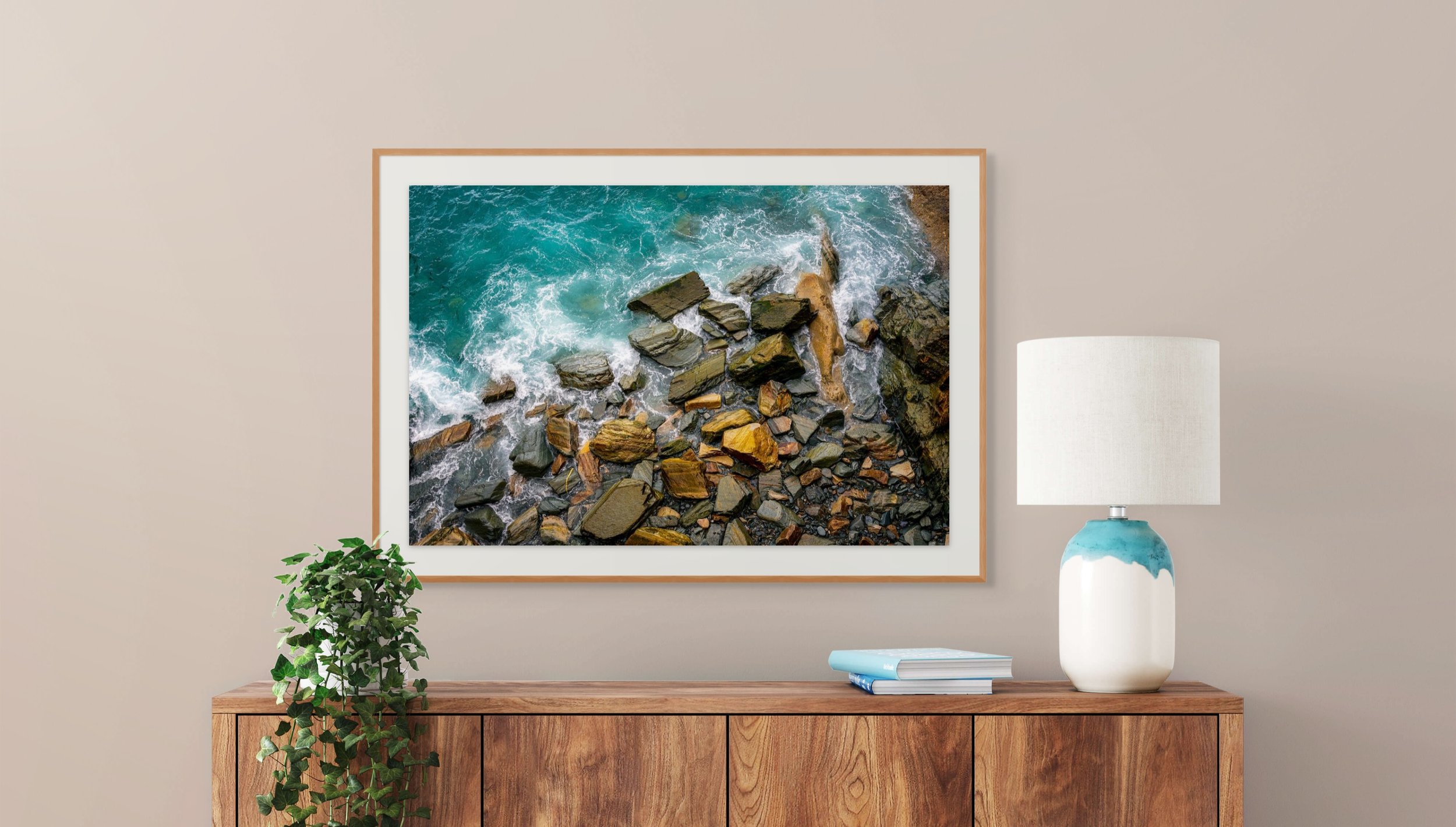 Framed artwork of a rocky shoreline with waves crashing against the rocks, hanging on a beige wall. A wooden sideboard below with a green potted plant on the left, a white and blue vase on the right, and some books.
