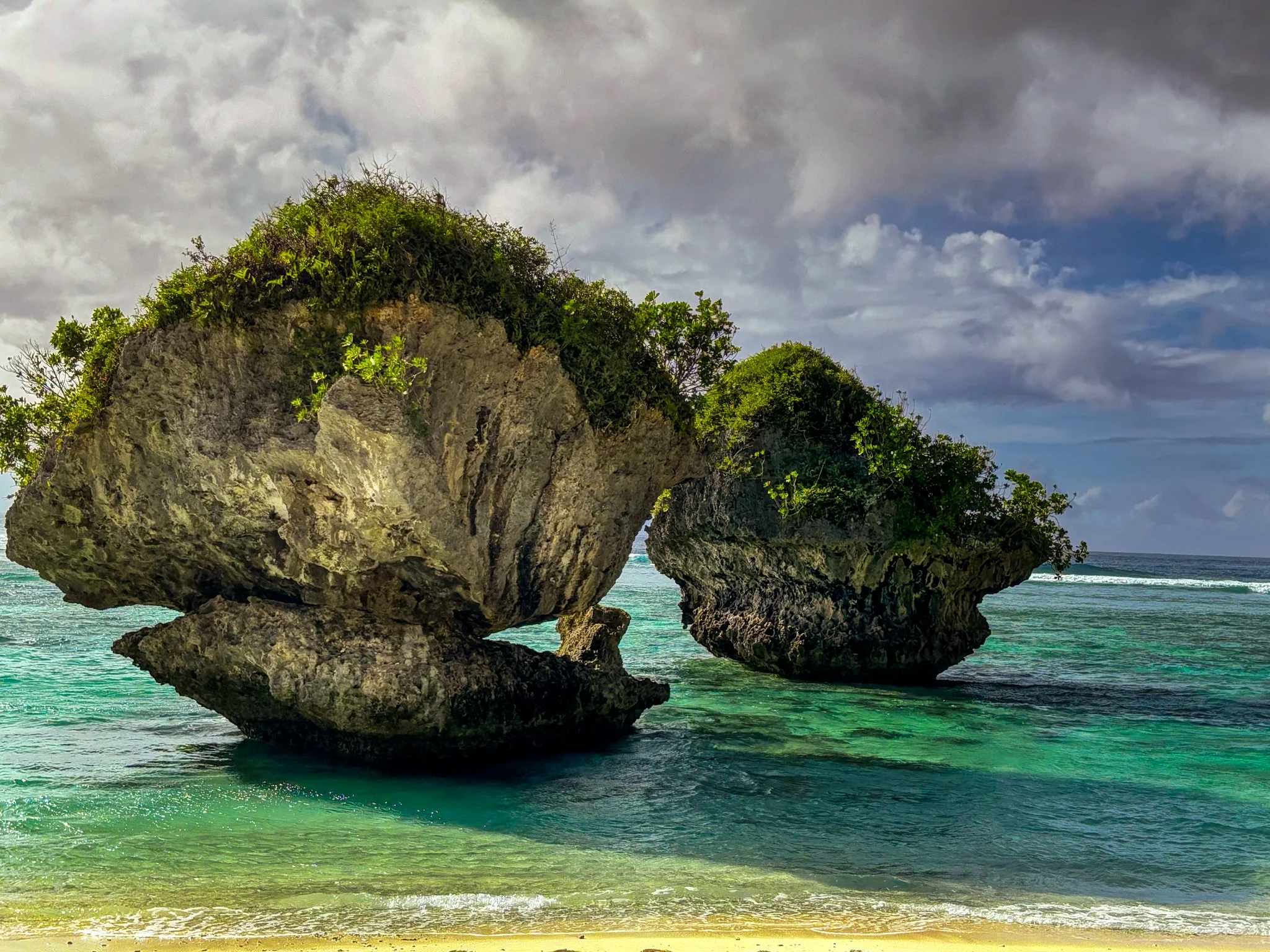 Large rock formations with green vegetation standing in shallow clear turquoise ocean water under a cloudy sky.