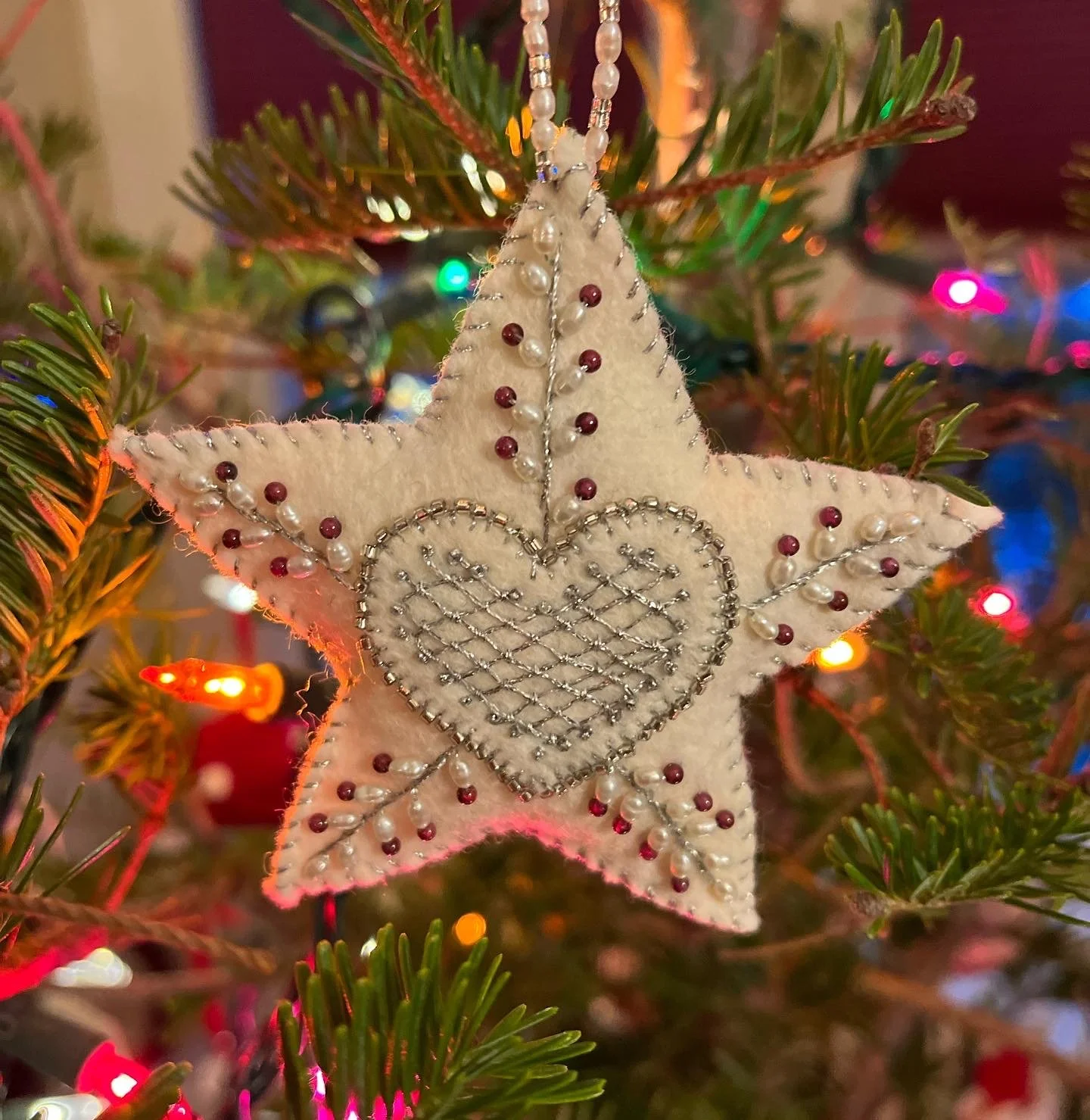 A star-shaped Christmas ornament decorated with a silver heart outline, pearls, and red beads hanging on a Christmas tree with colorful lights.