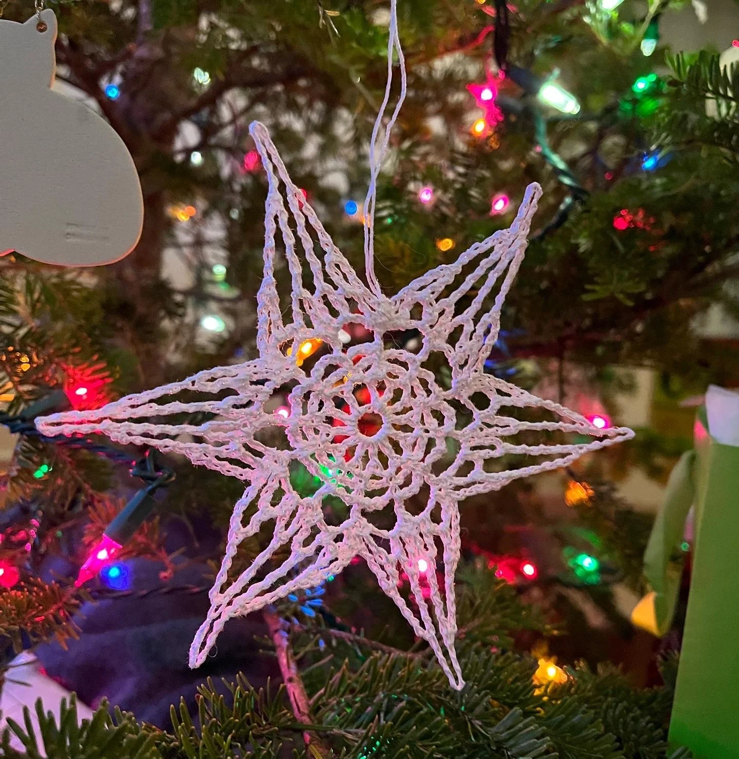 A white, three-dimensional star-shaped Christmas ornament hanging on a Christmas tree with multicolored lights.