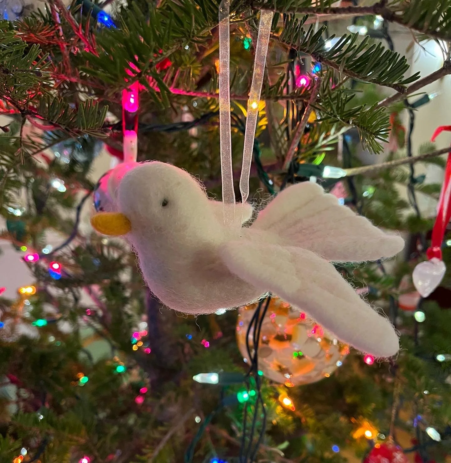 A plush white bird ornament with an orange beak hanging on a decorated Christmas tree with colorful lights.