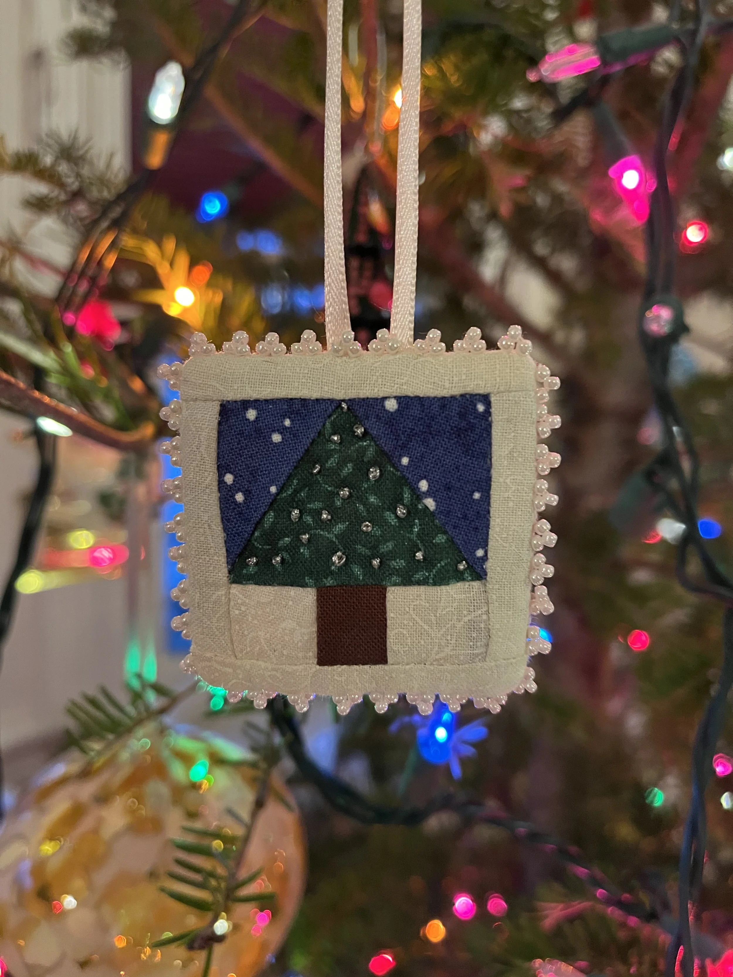 A small fabric Christmas ornament hanging from a Christmas tree, decorated with beads and featuring a green Christmas tree design. The background displays colorful blurred Christmas lights.
