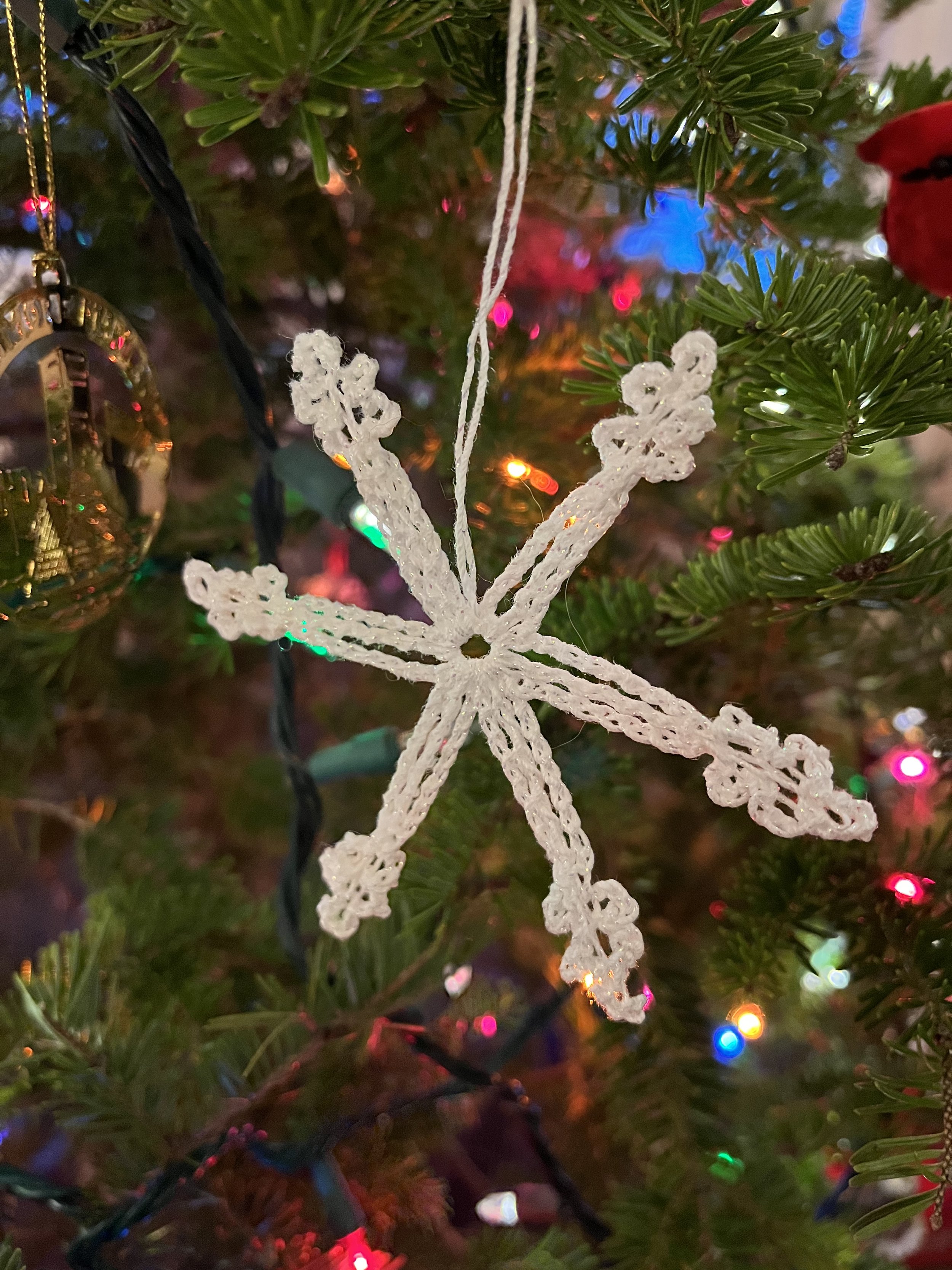 A close-up of a white snowflake-shaped ornament hanging on a Christmas tree. The background shows colorful Christmas lights.