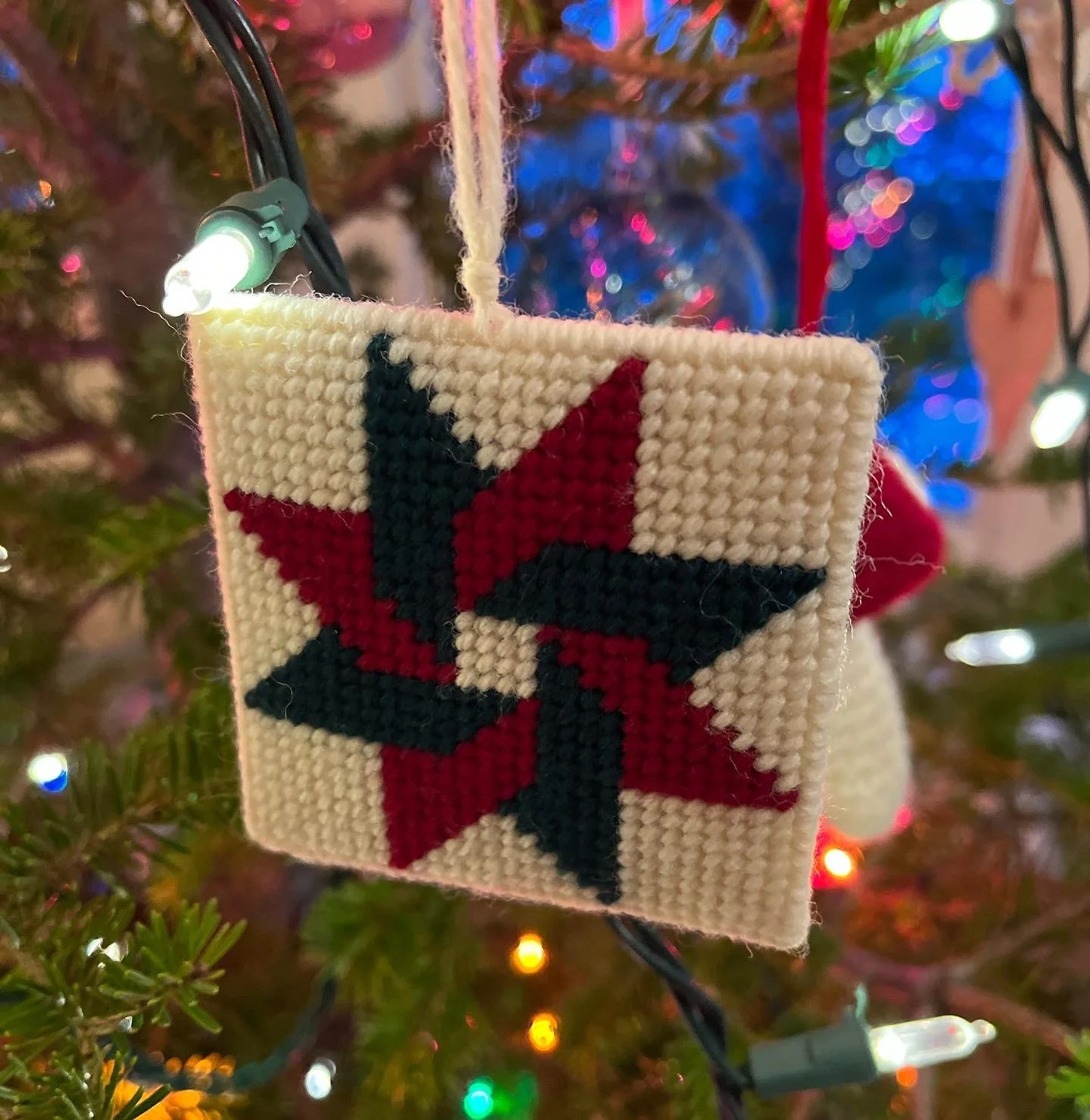 Close-up of a Christmas ornament with a black and red pinwheel design, hanging on a decorated Christmas tree with colorful lights in the background.