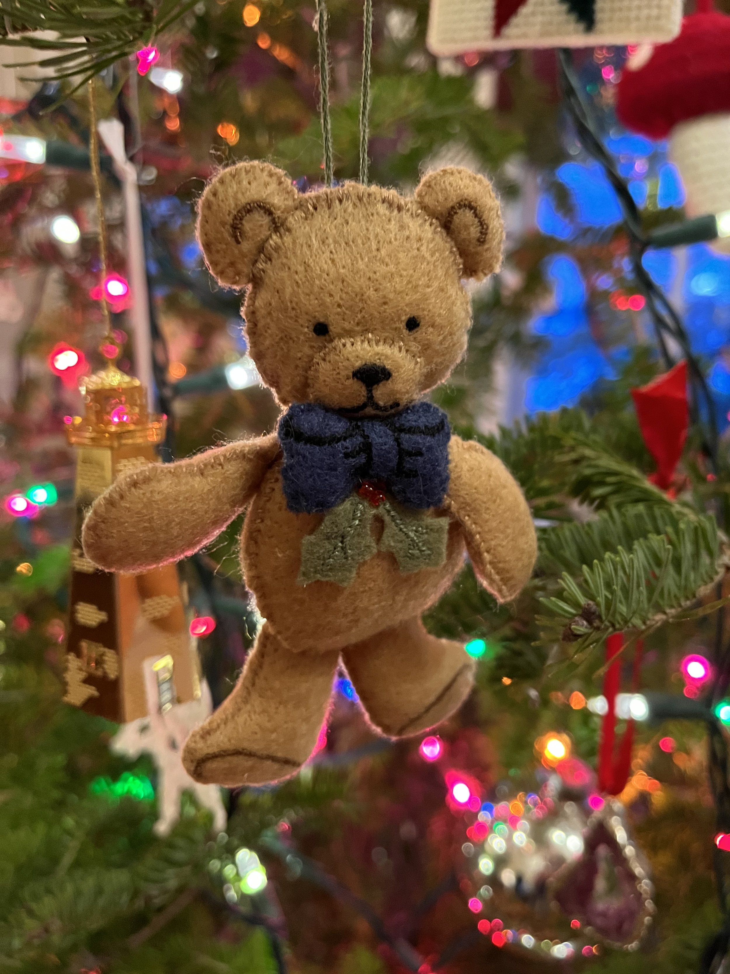 A small teddy bear Christmas ornament hanging on a decorated Christmas tree with colorful lights in the background.