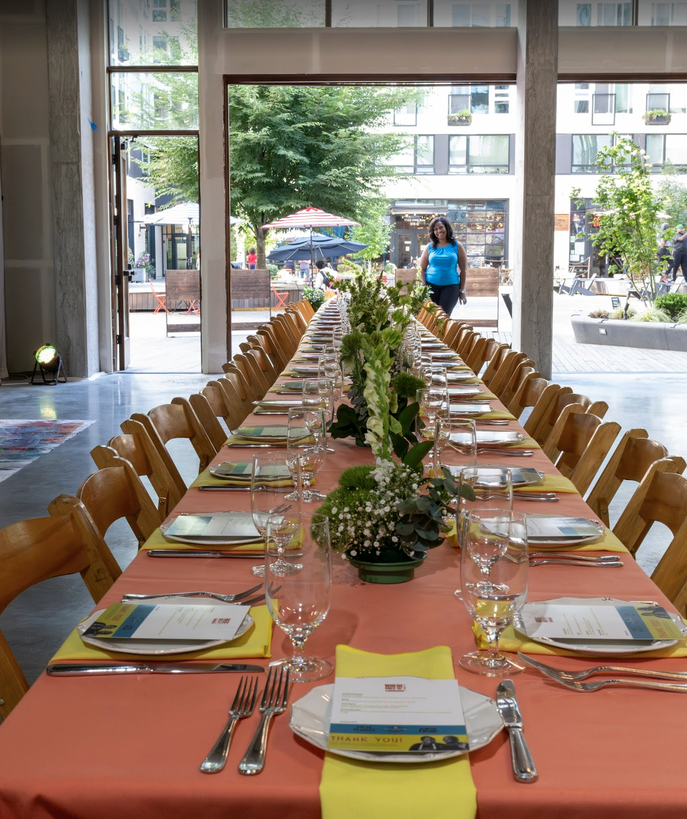 A long dining table set for a celebration or event with peach tablecloths, yellow napkins, and place settings including silverware, glasses, and menus. The table is decorated with white and green floral centerpieces. Large glass doors open to an outdoor patio with trees, umbrellas, and people walking by. A woman in a blue top is outside, smiling at the camera.