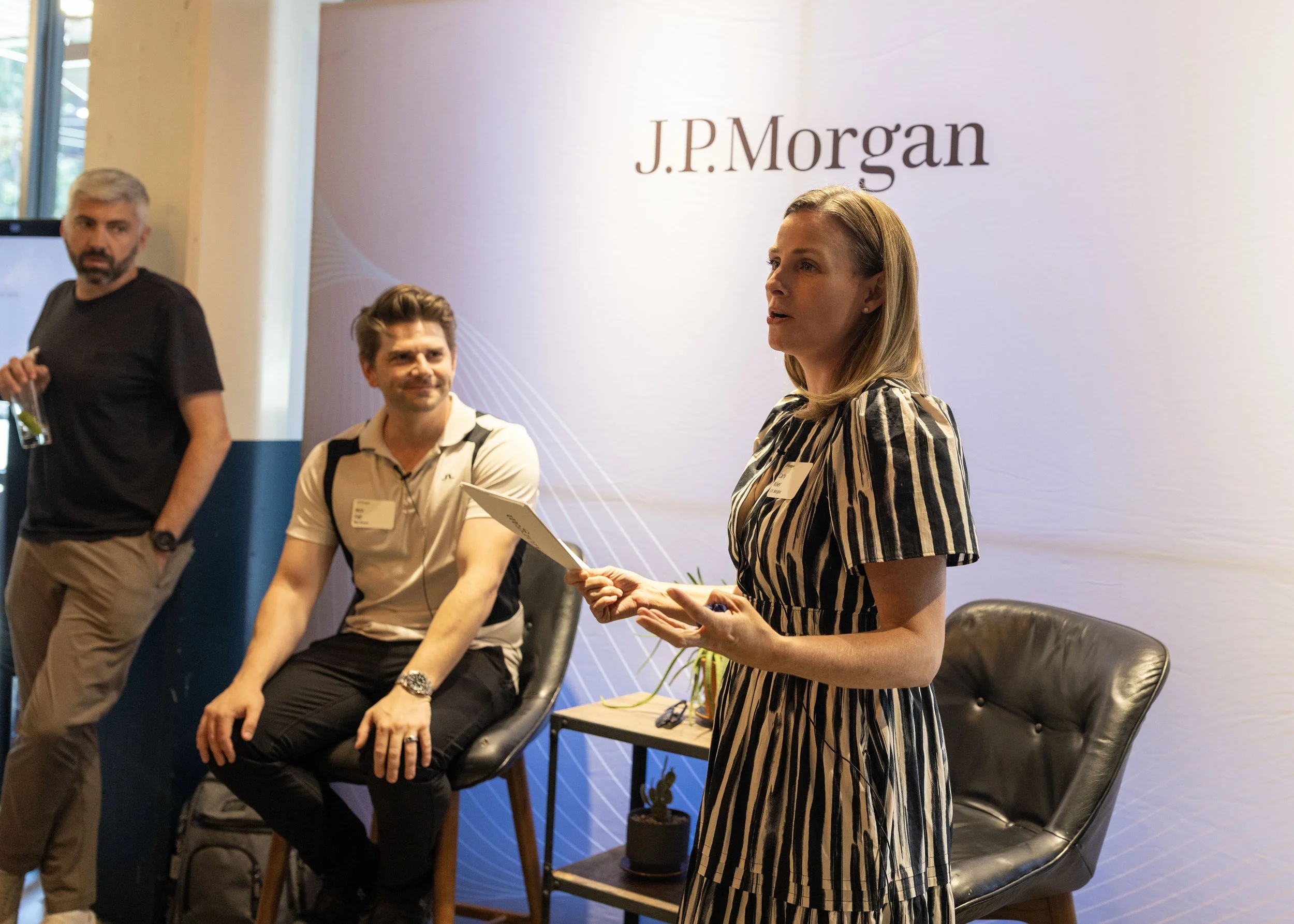 A woman with blonde hair speaking at an event with a J.P. Morgan sign in the background. Two men are seated beside her, one with a beard and the other with brown hair, listening attentively.