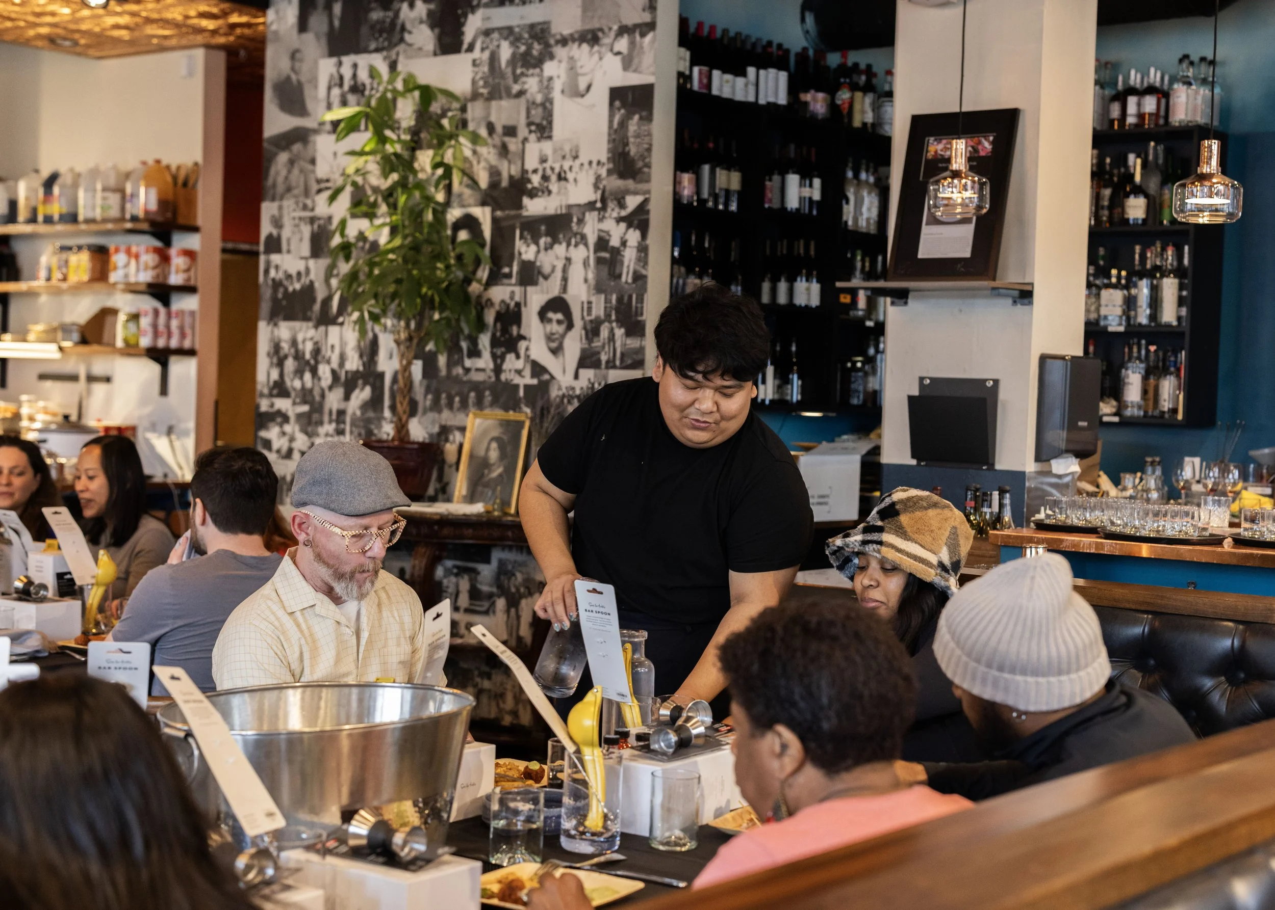 A group of people dining at a restaurant, some sitting at a long table and others standing. The scene features a background with shelves of bottles and a collage of black and white photos on the wall. The atmosphere appears lively and social, with a man in a black shirt serving or assisting customers.