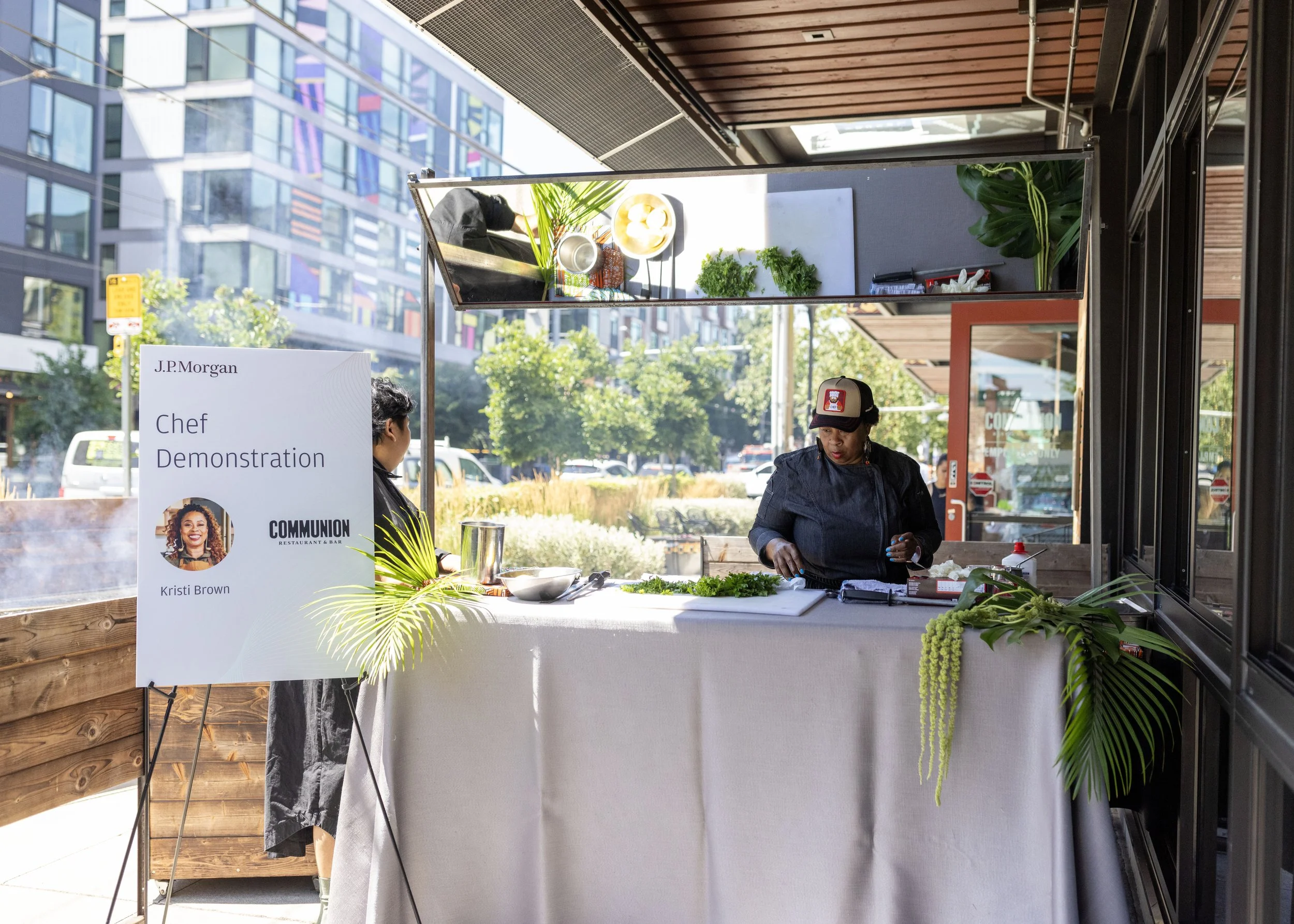 A woman cooking or preparing food at a demonstration stand outdoors, with a sign that reads "J.P. Morgan Chef Demonstration Kristi Brown," and various plants and cooking utensils on the table.
