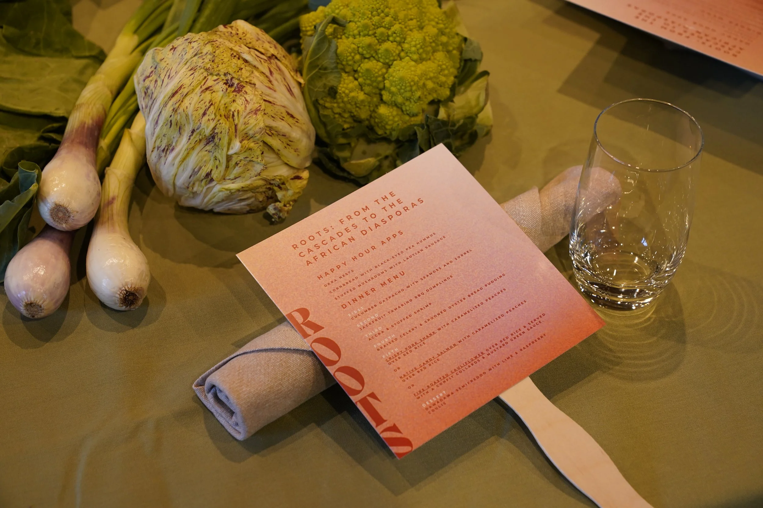 Fresh vegetables including garlic, cauliflower, broccoli, and what appears to be yellow Romanesco, arranged on a table near you a pink menu and a glass of water.