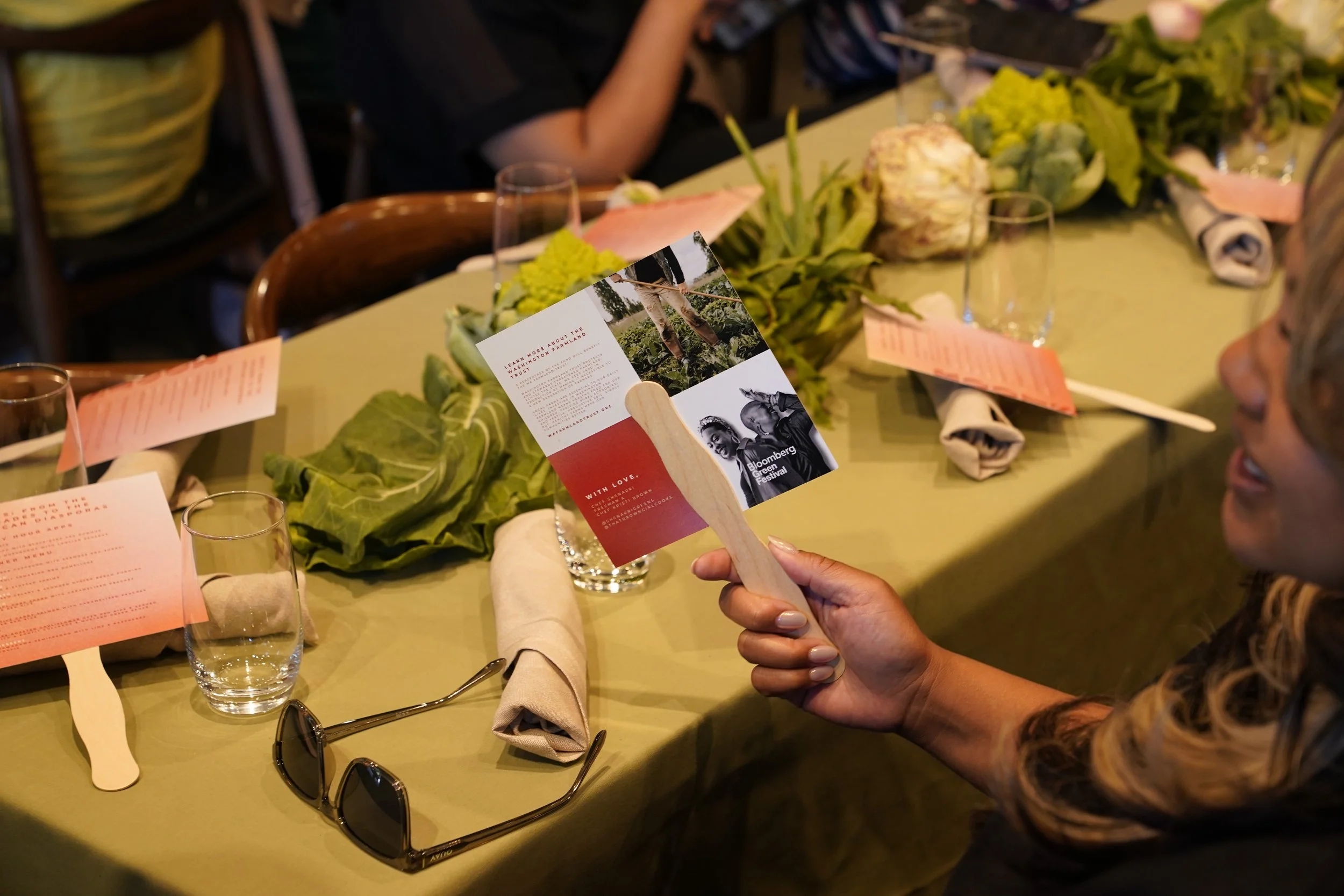 A woman holding a wooden card with printed text and images, seated at a table with a green tablecloth, surrounded by glasses, napkins, and floral decorations, during a social event or dinner.