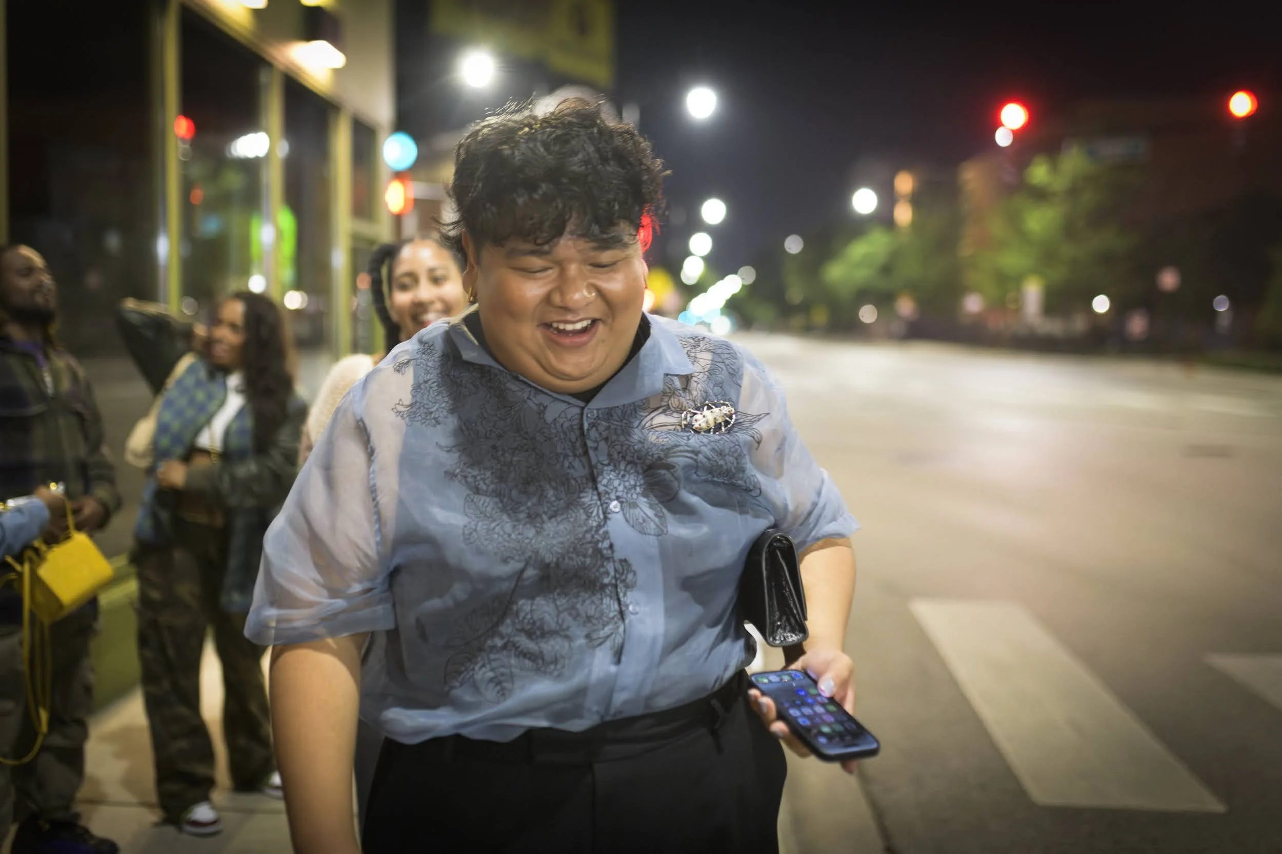A group of people standing on a city sidewalk at night, with a young man in the foreground smiling and looking at his phone, others behind him chatting and smiling, illuminated by streetlights and traffic signals.
