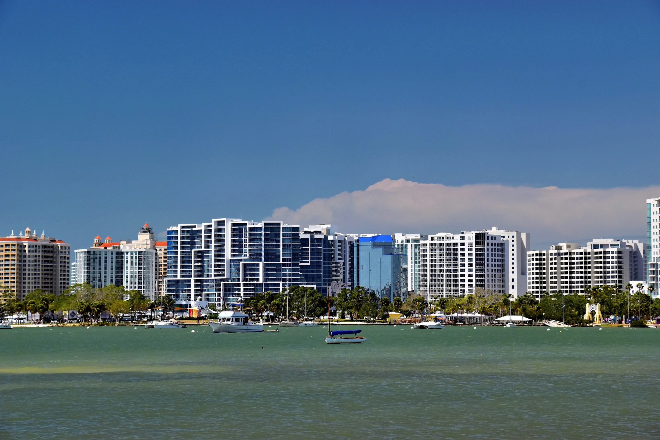 City skyline with modern high-rise buildings along the waterfront, boats anchored in the water in the foreground, and a clear blue sky with some clouds.