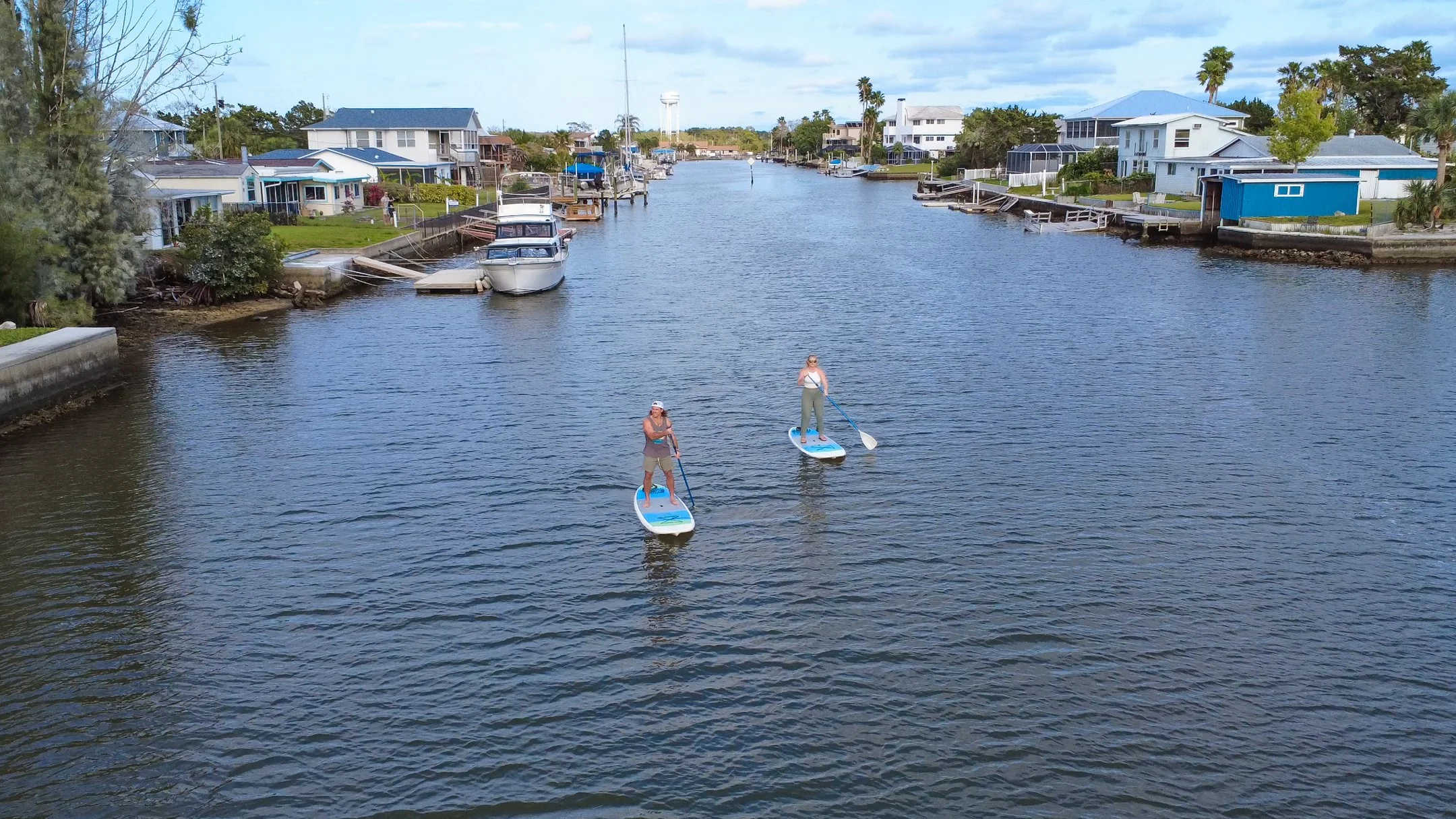 Two people paddleboarding in a narrow waterway lined with houses and boats, with a water tower in the background.