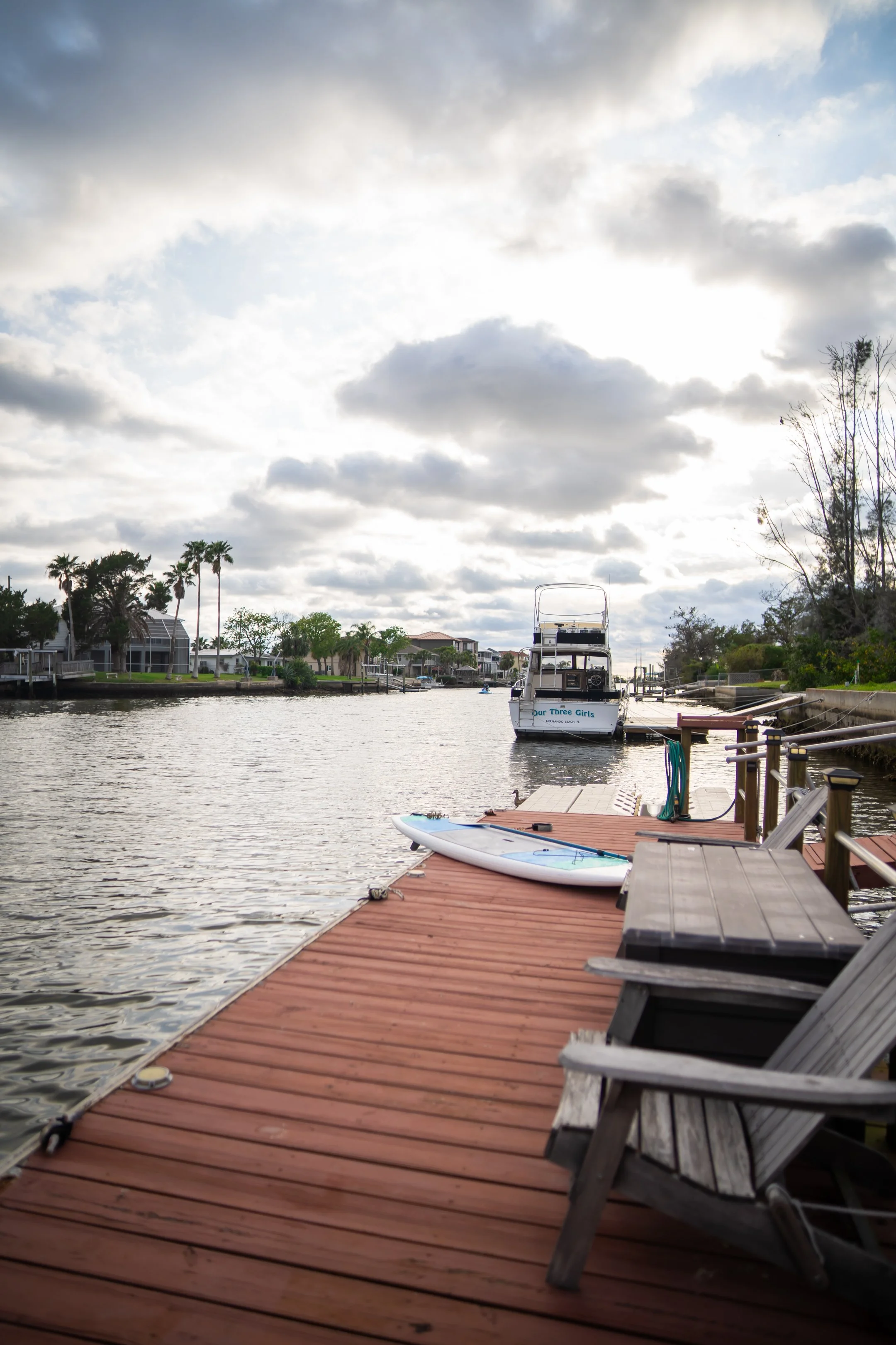A view of a canal or river with houses and palm trees along the shoreline, boats docked or sailing, and a mostly cloudy sky overhead.