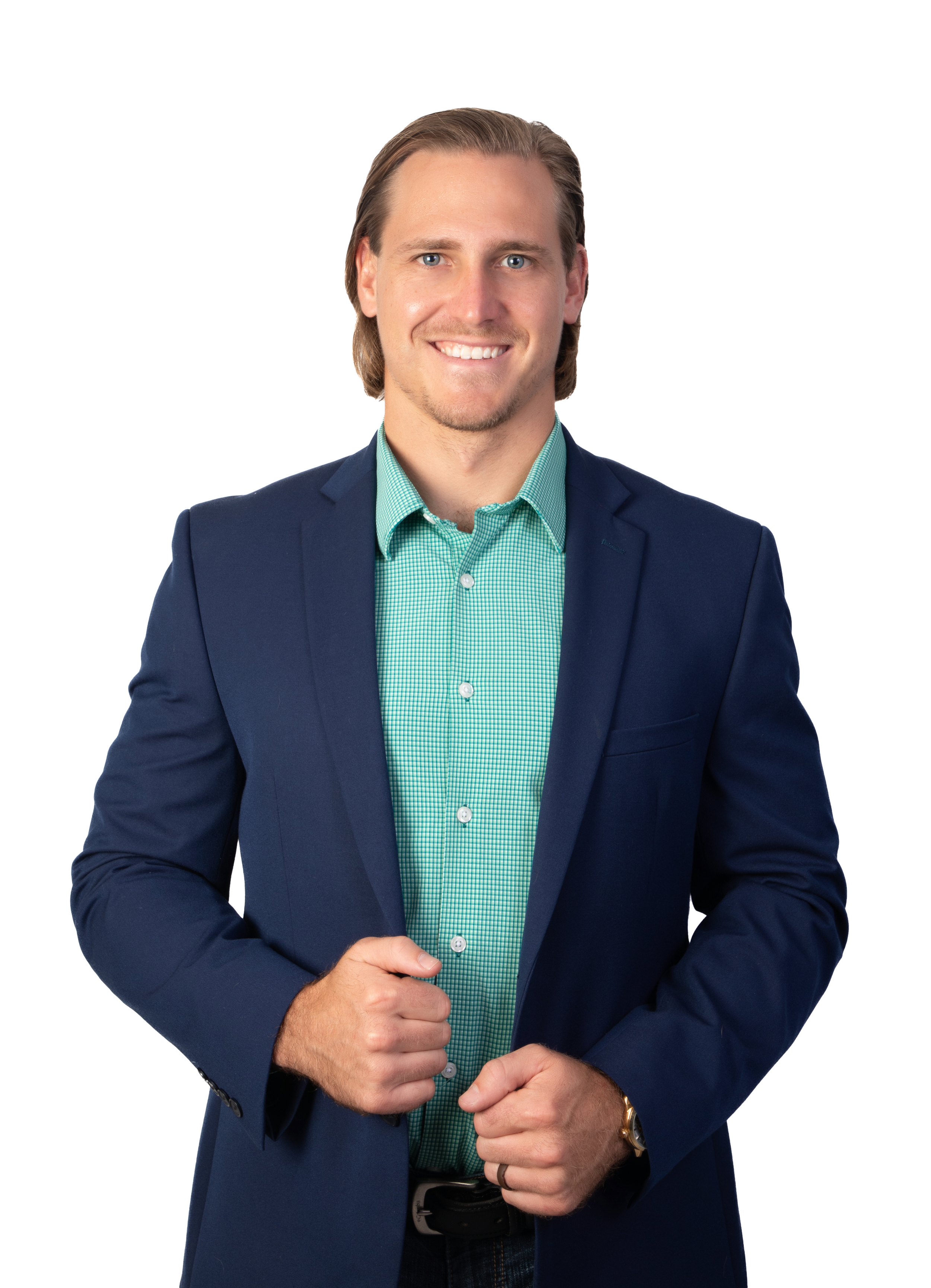 A man with light skin, brown hair, and blue eyes, wearing a blue blazer and a green checkered shirt, smiling confidently with his hands clasped in front of him.
