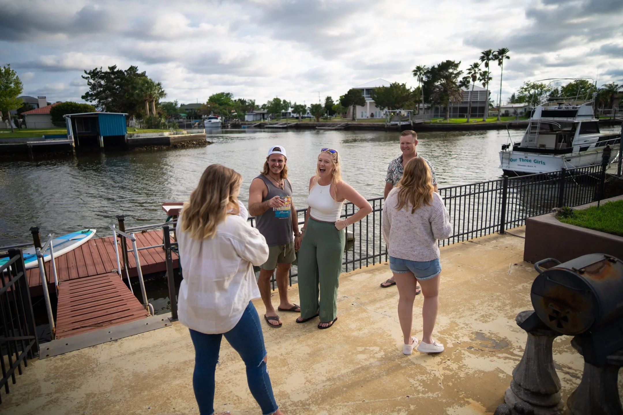 A group of six people standing on a dock near the water, laughing and talking, with boats and houses in the background, under a partly cloudy sky.