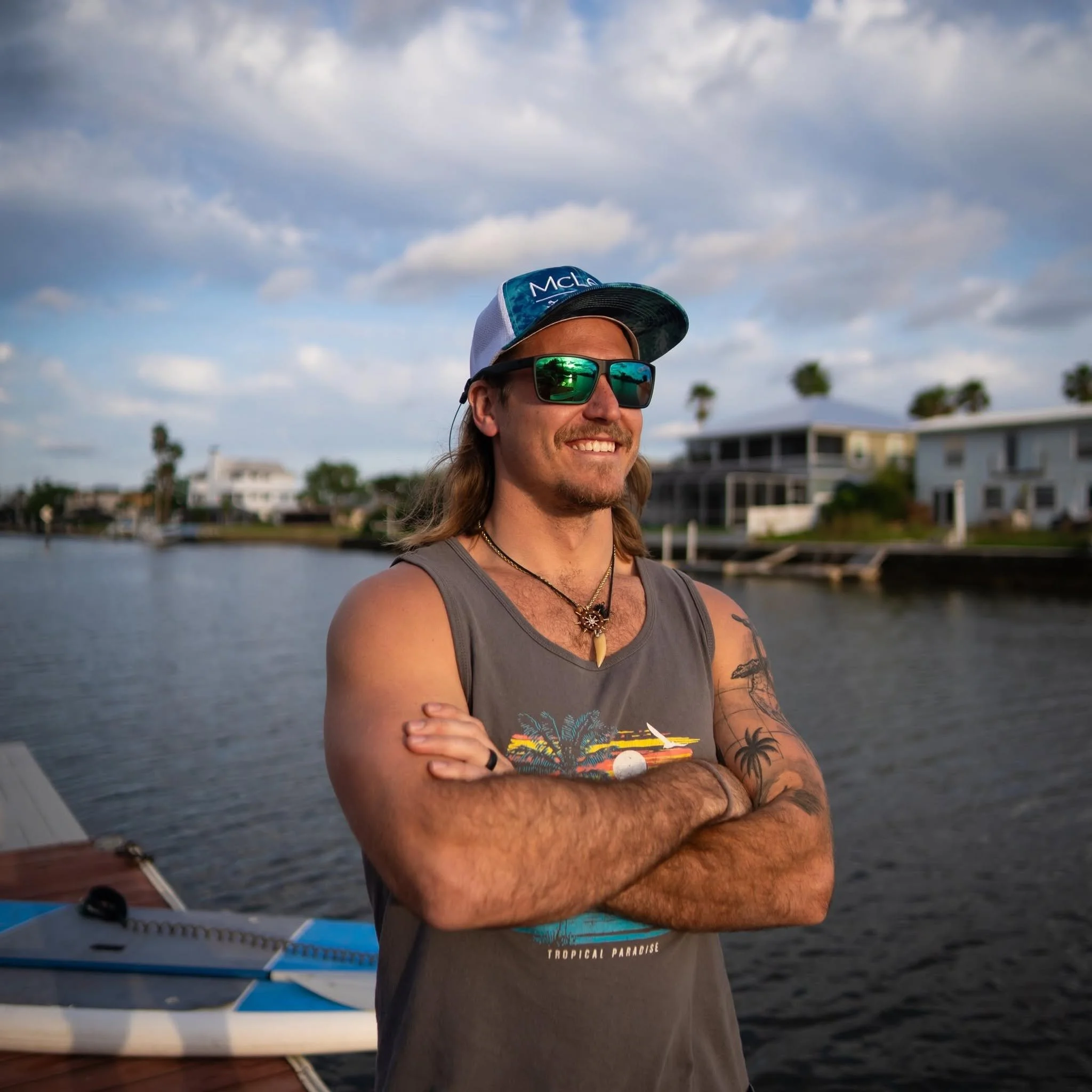 A person with long hair, wearing sunglasses, a cap, and a tank top, standing outdoors near water with boats and buildings in the background.