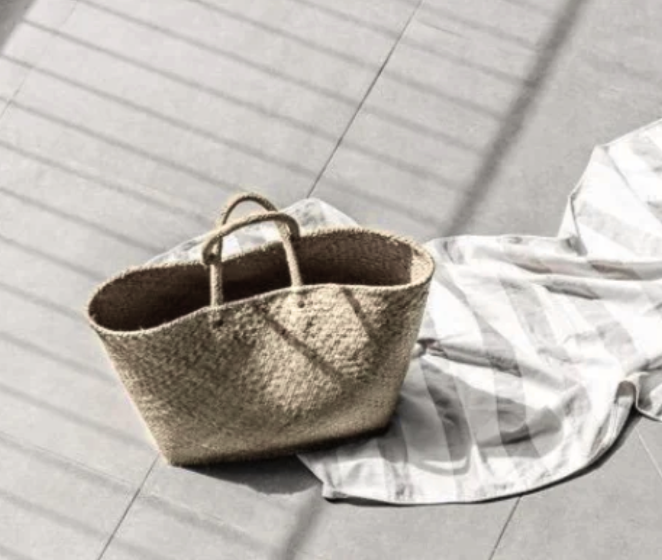 A beige woven tote bag with handles on a tiled floor, partially resting on a white cloth with light gray stripes.