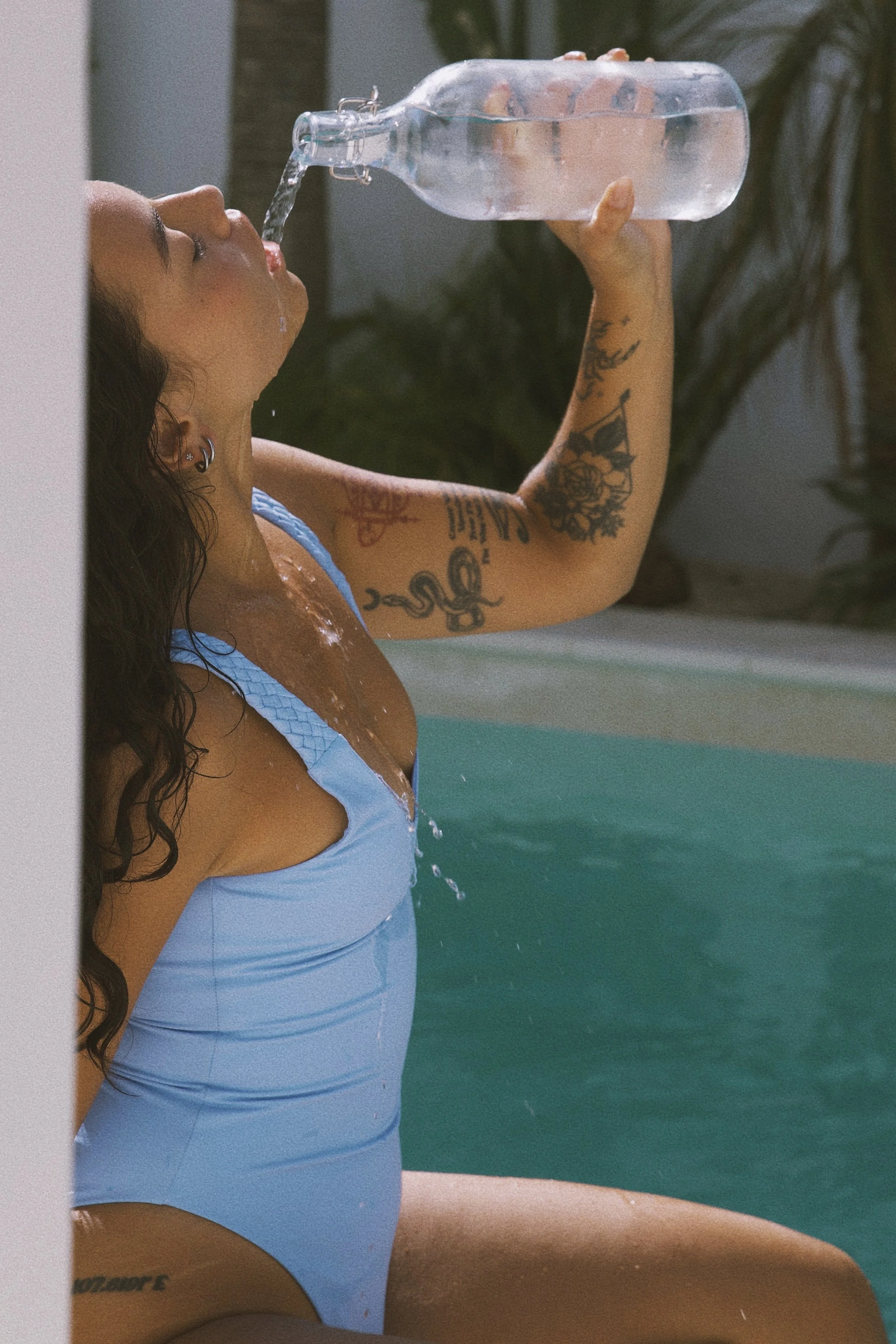 A woman with long dark hair, tattoos on her arm, and earrings is playing with a bottle of water, pouring it into her mouth while sitting by a pool. She is wearing a light blue swimsuit.